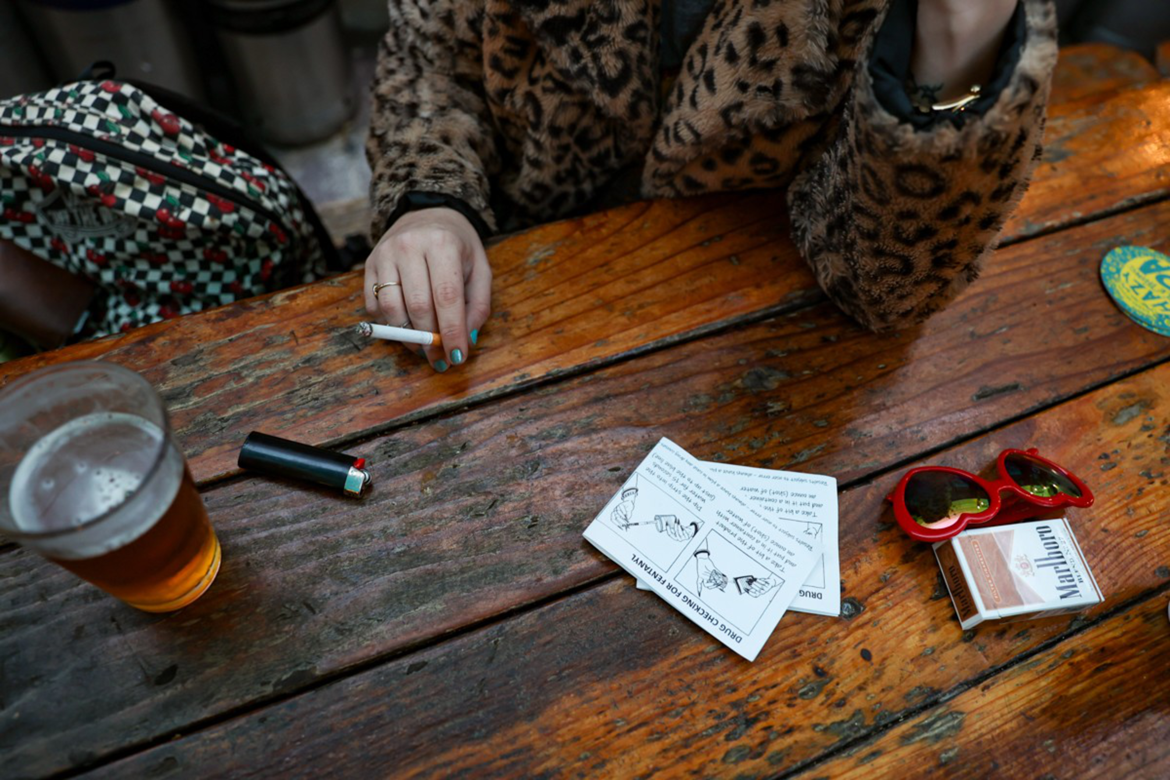 A person in a leopard print jacket holds a cigarette at a wooden table with a lighter, beer, red sunglasses, Marlboro pack, and paper instructions.