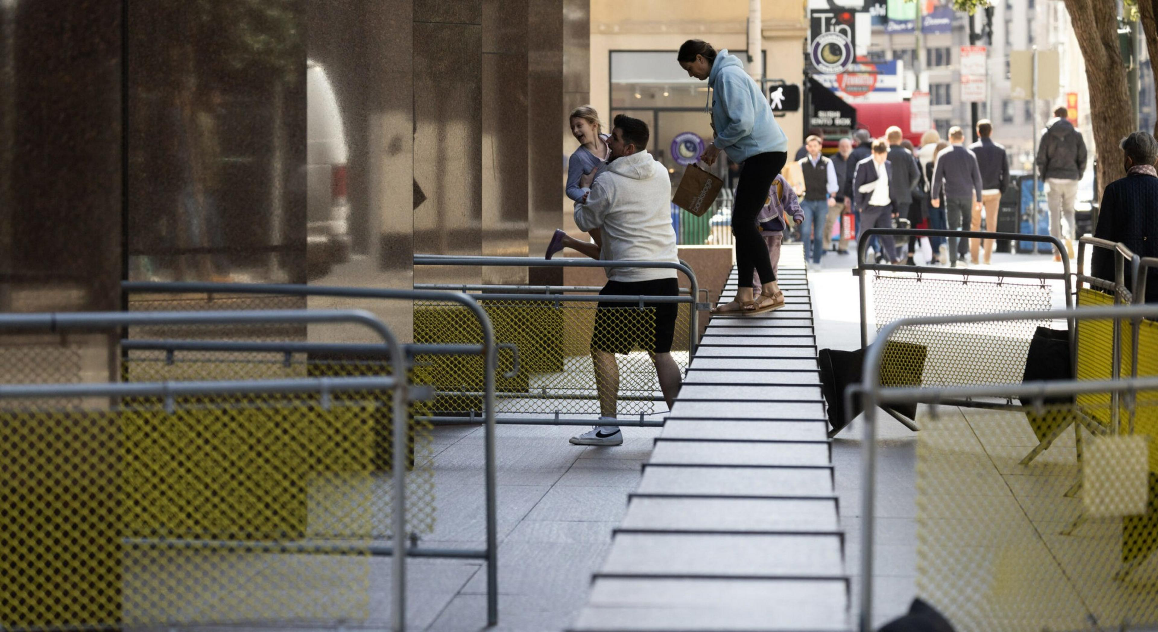 A man holds a child while a woman stands nearby on a narrow elevated walkway in a city area with pedestrians walking in the background.