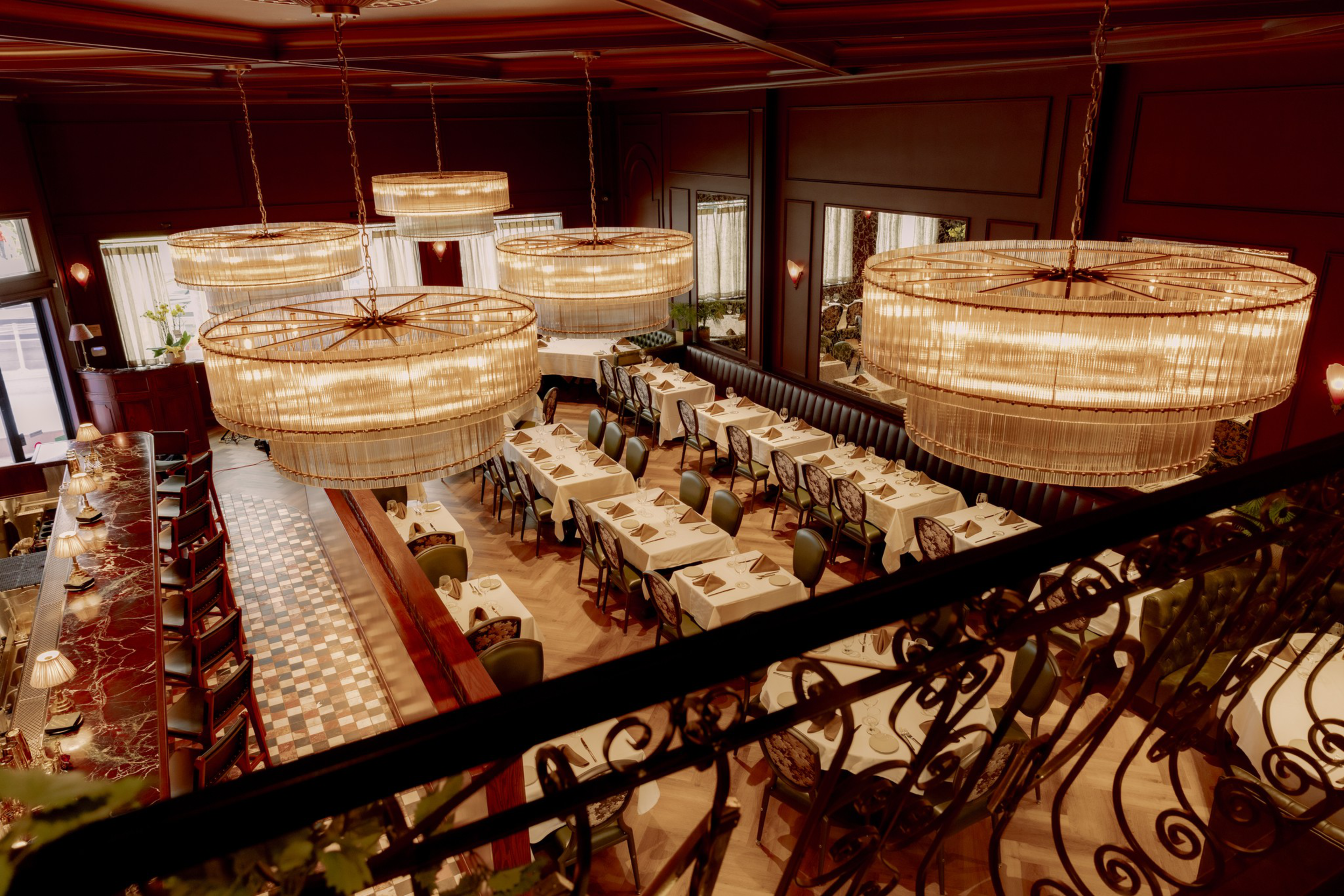 Elegant restaurant dining area with white tablecloths, green cushioned chairs, large circular chandeliers, and a polished bar with red marble counters.