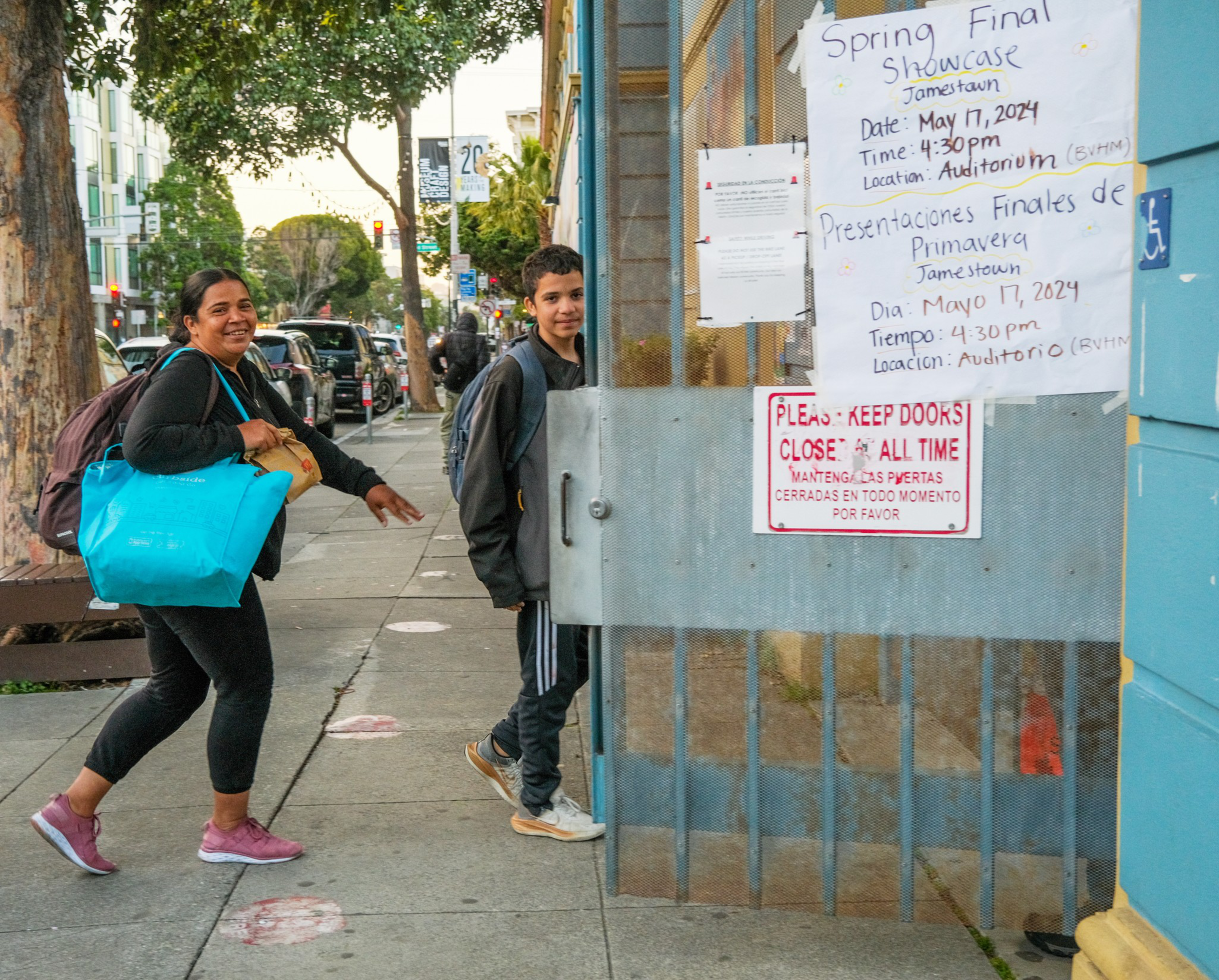 A woman and a boy stand by a gate with event posters; urban background.