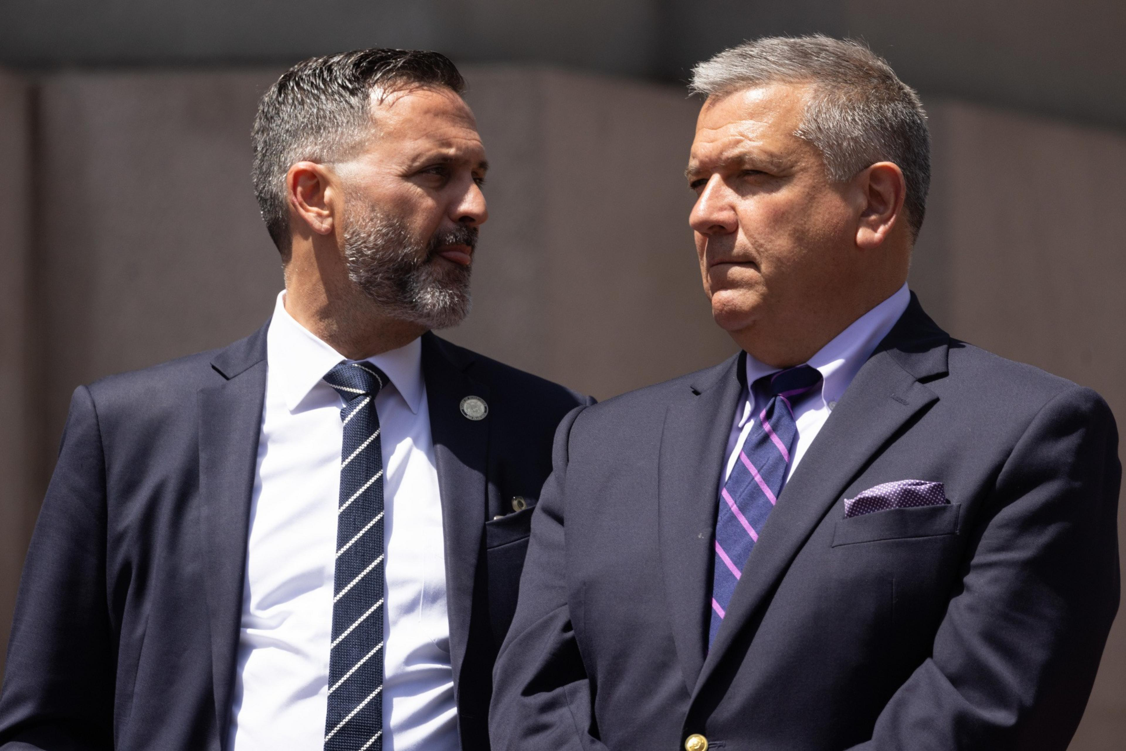 Two men in suits are standing together, one looking to the side and the other straight ahead.