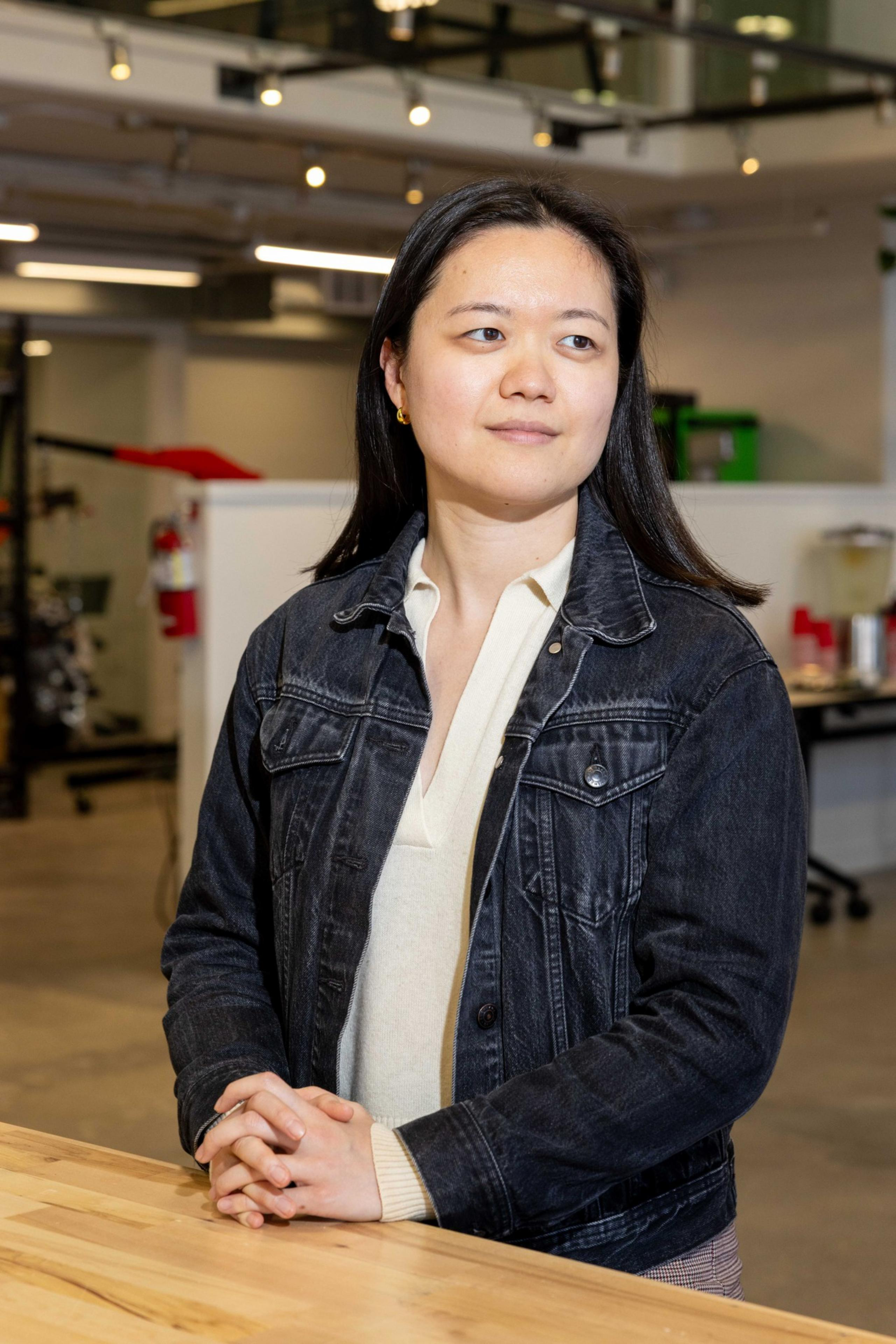 A person with long dark hair is standing indoors, wearing a denim jacket over a light shirt, with hands clasped on a wooden table. The background is an industrial setting.