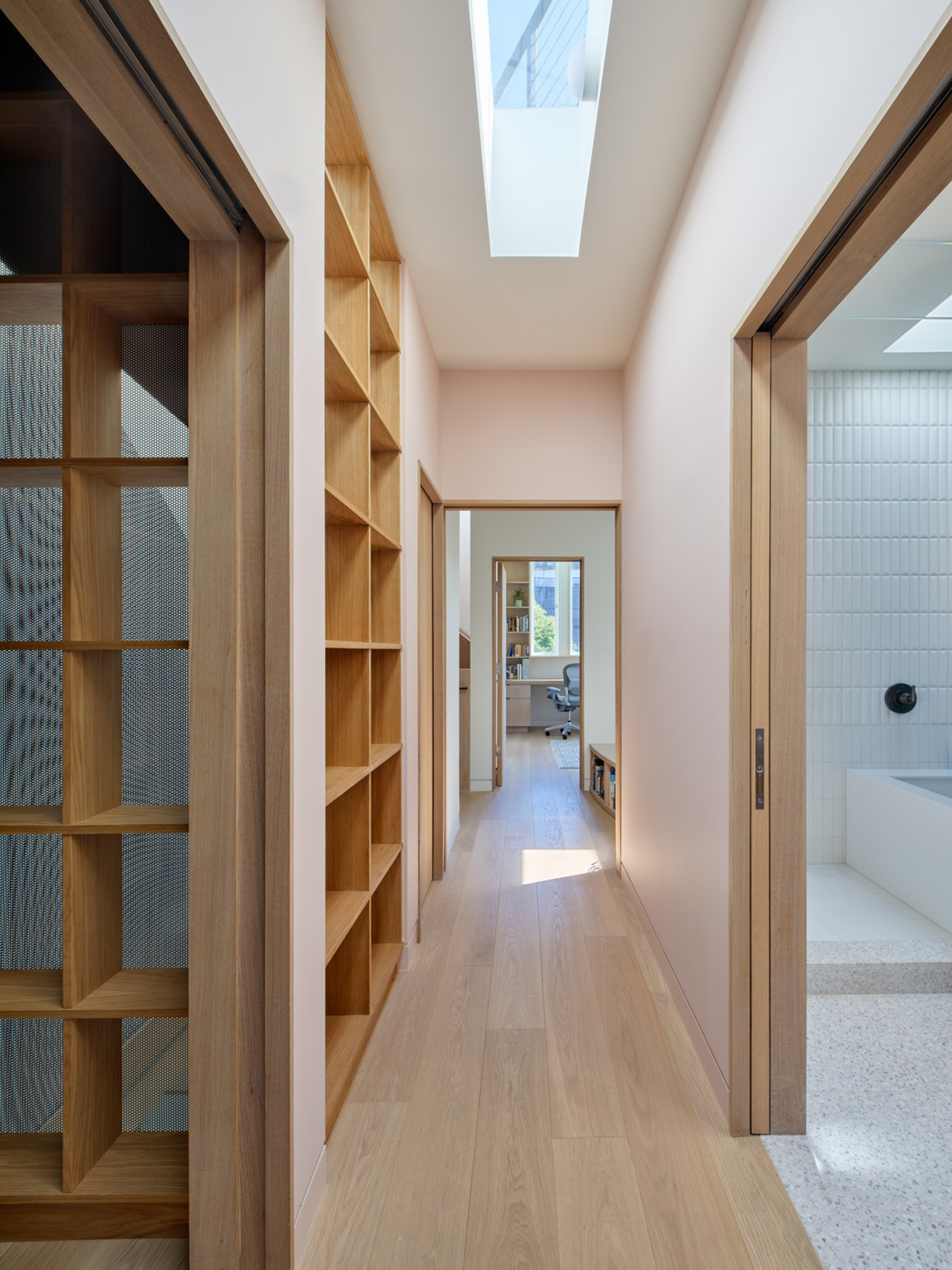 A bright hallway with light wood floors and shelves on the left, skylight above, sliding wooden doors on both sides, leading to an office room.