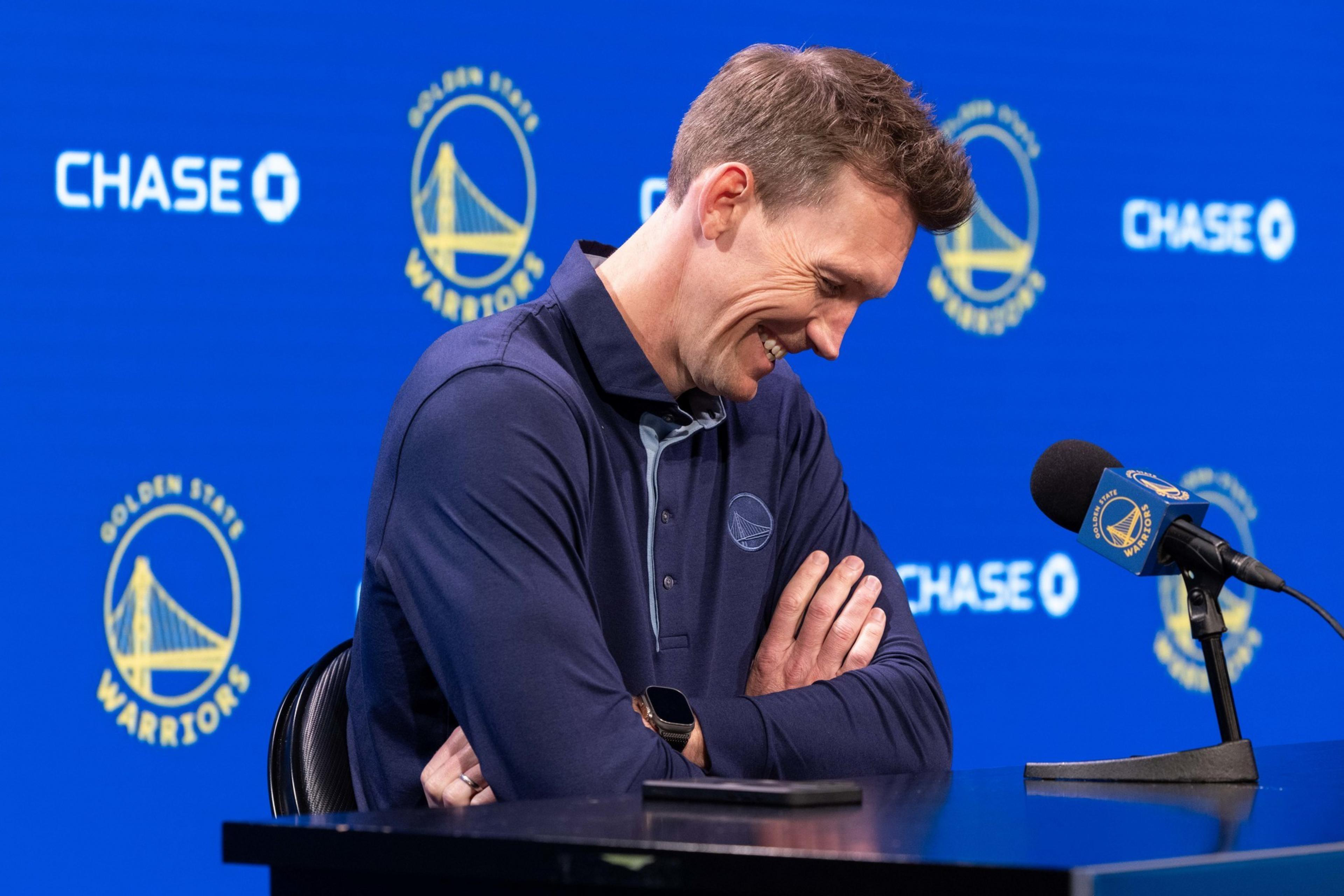 A smiling man stands at a microphone, arms crossed, in front of a blue background with Golden State Warriors logos and the word &quot;CHASE&quot; visible.