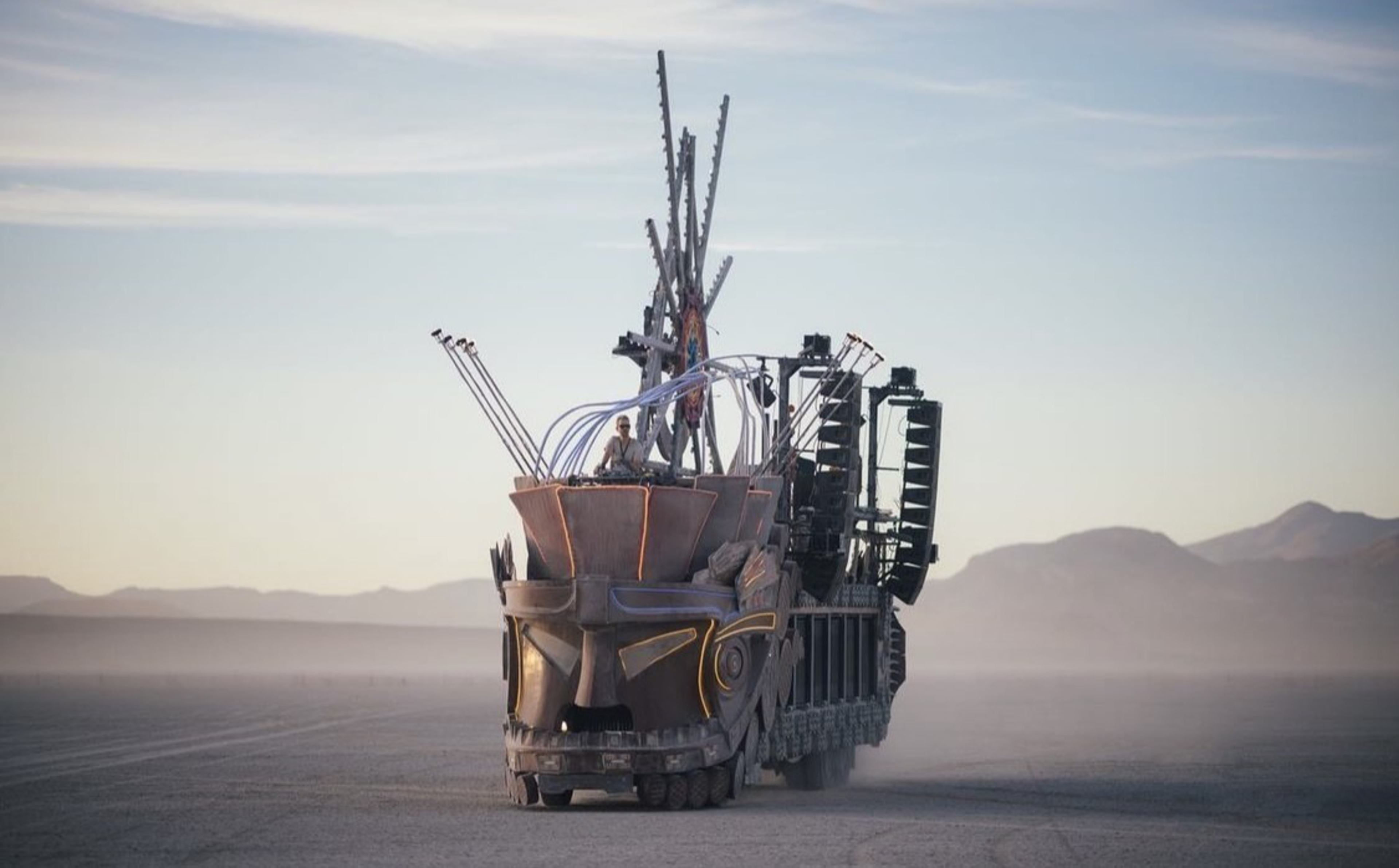 A large, intricately designed vehicle resembling a tribal mask with tall structures and speakers moves across a flat desert landscape.