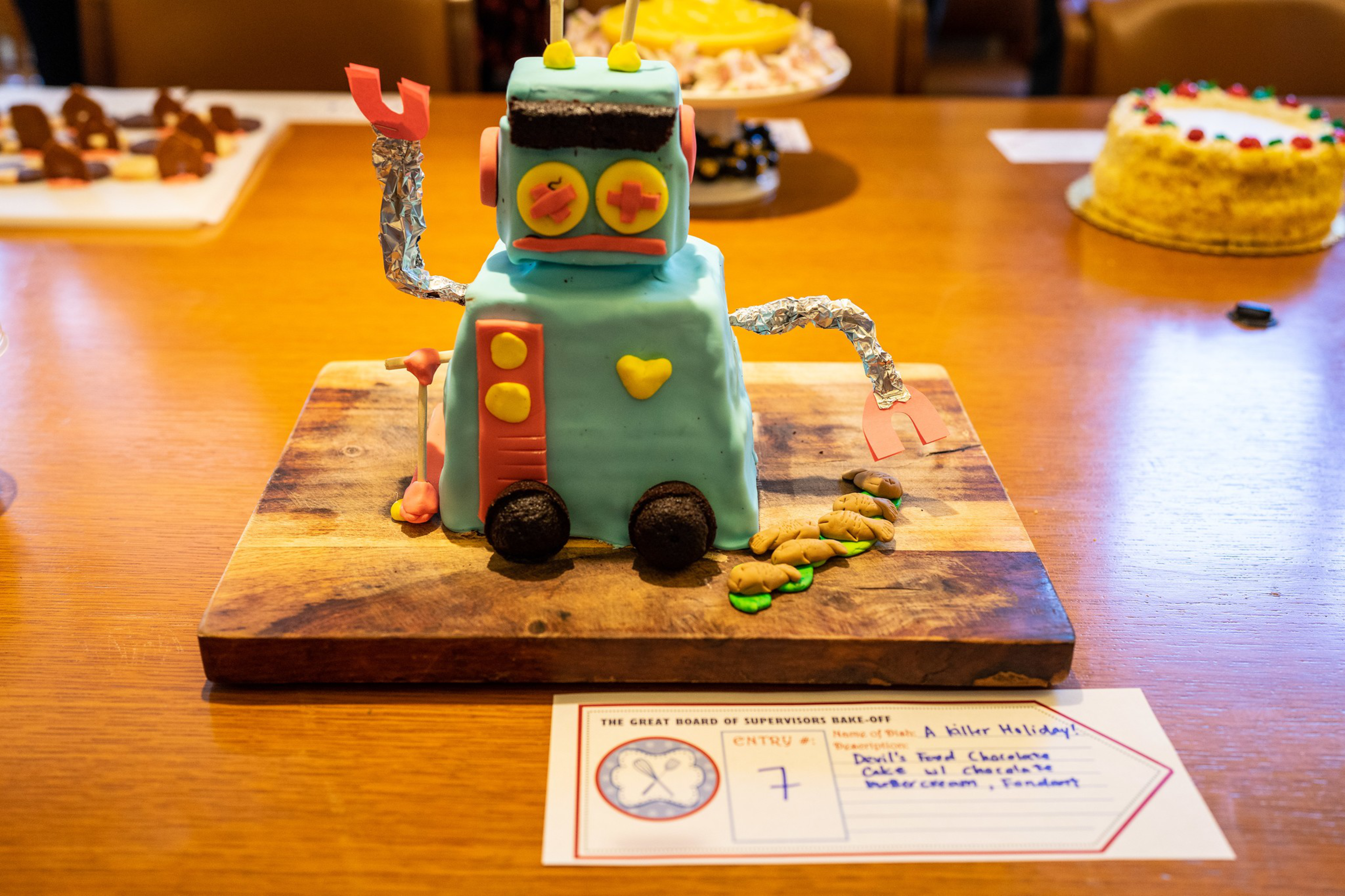A robot-shaped cake covered in blue fondant with foil arms and candy eyes sits on a wooden board, surrounded by other cakes on a table.
