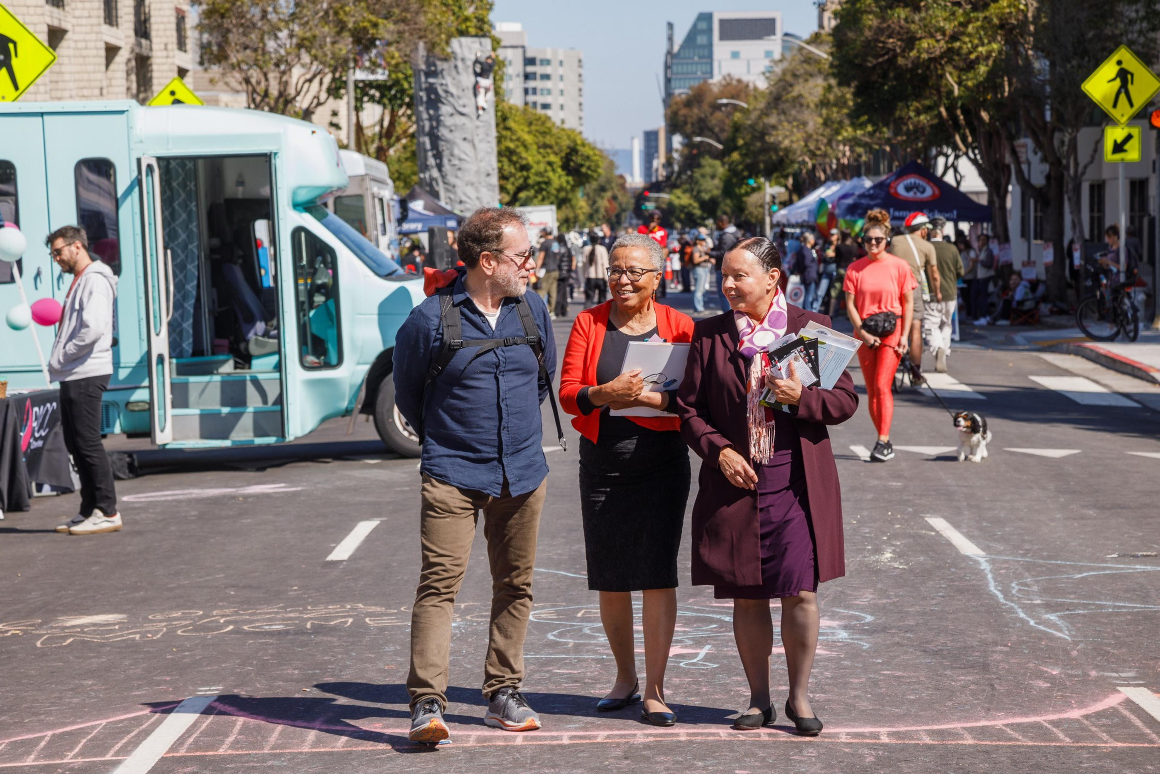 Three people chat on a bustling street with chalk art, a teal bus, climbing wall, and passersby in the background.