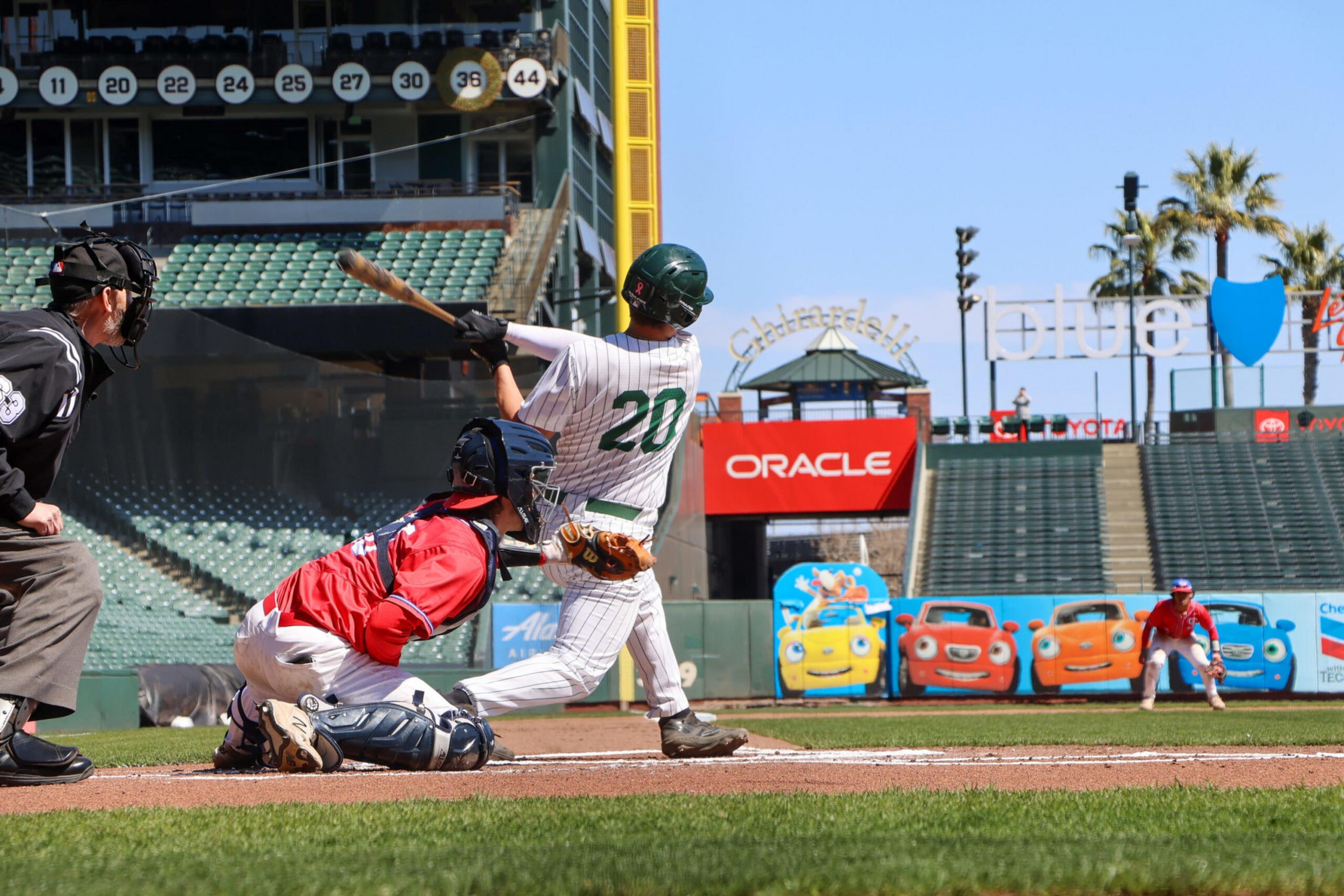 A baseball player in a white pinstripe uniform swings at a pitch while the catcher in red crouches behind home plate and the umpire watches closely.