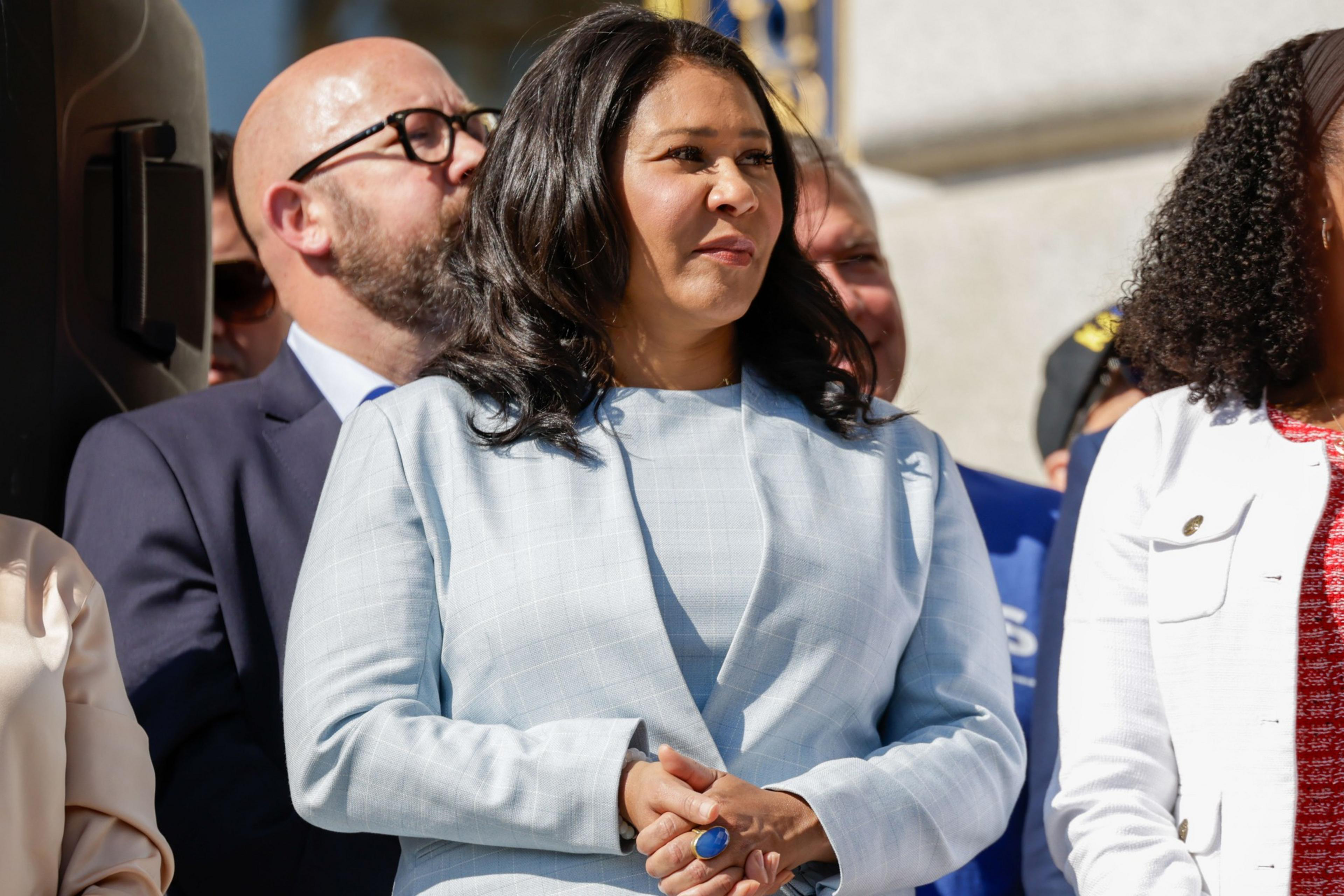 A woman in a light blue blazer stands outdoors among a group of people, some in suits. She has dark hair and appears to be listening attentively.