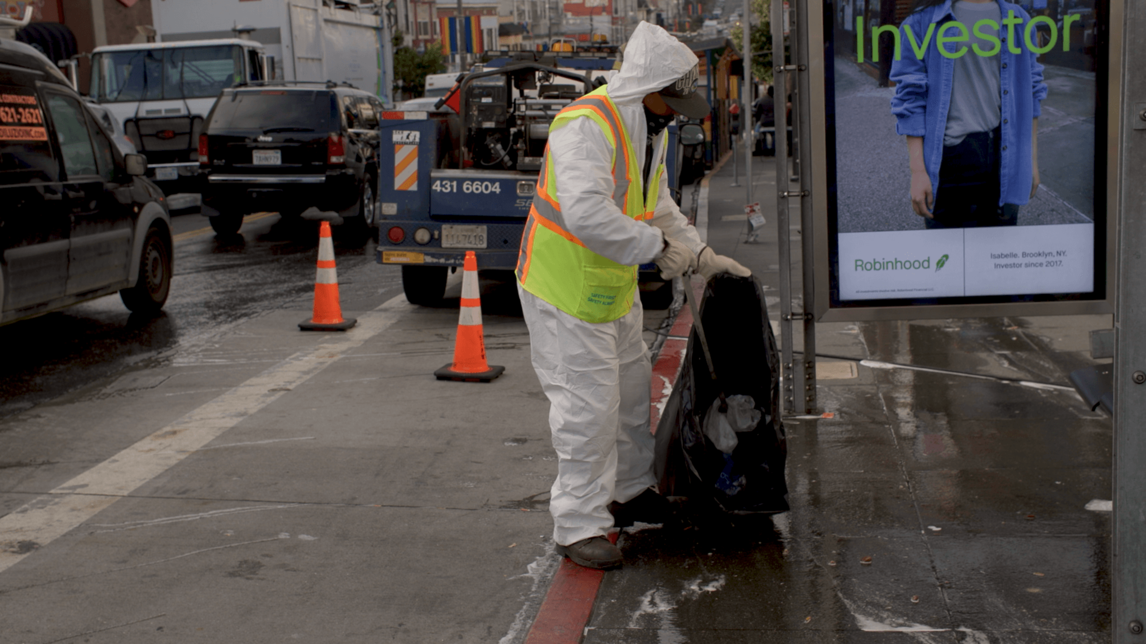 A person wearing a white protective suit and a yellow safety vest picks up trash from the curb near a bus stop on a wet street.