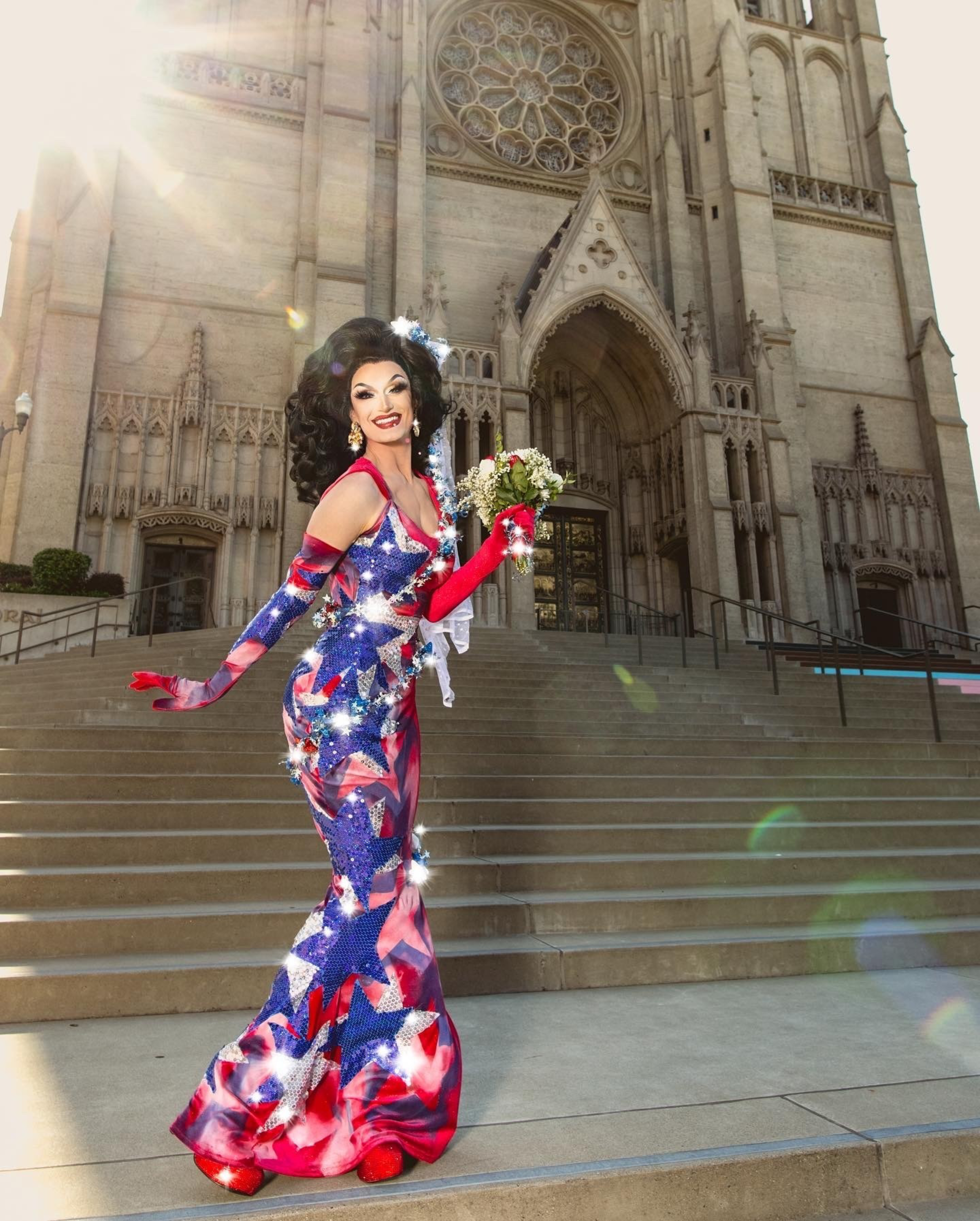 A person in a vibrant, glittery red, white, and blue gown with matching gloves poses joyfully on church steps, holding a small bouquet of flowers.