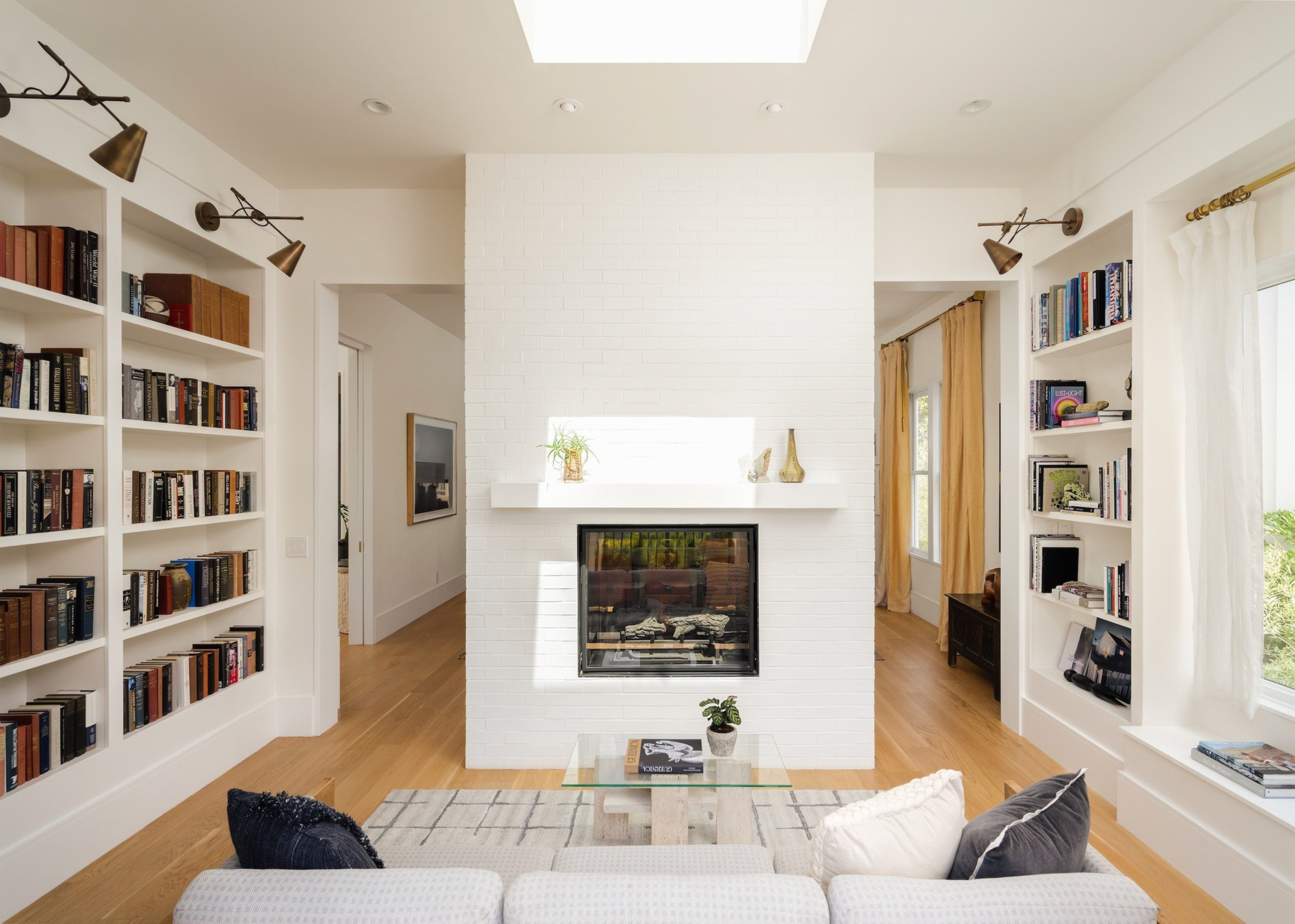 A bright living room features a white brick fireplace, built-in bookshelves filled with books, a glass coffee table, and a light sofa with dark pillows.