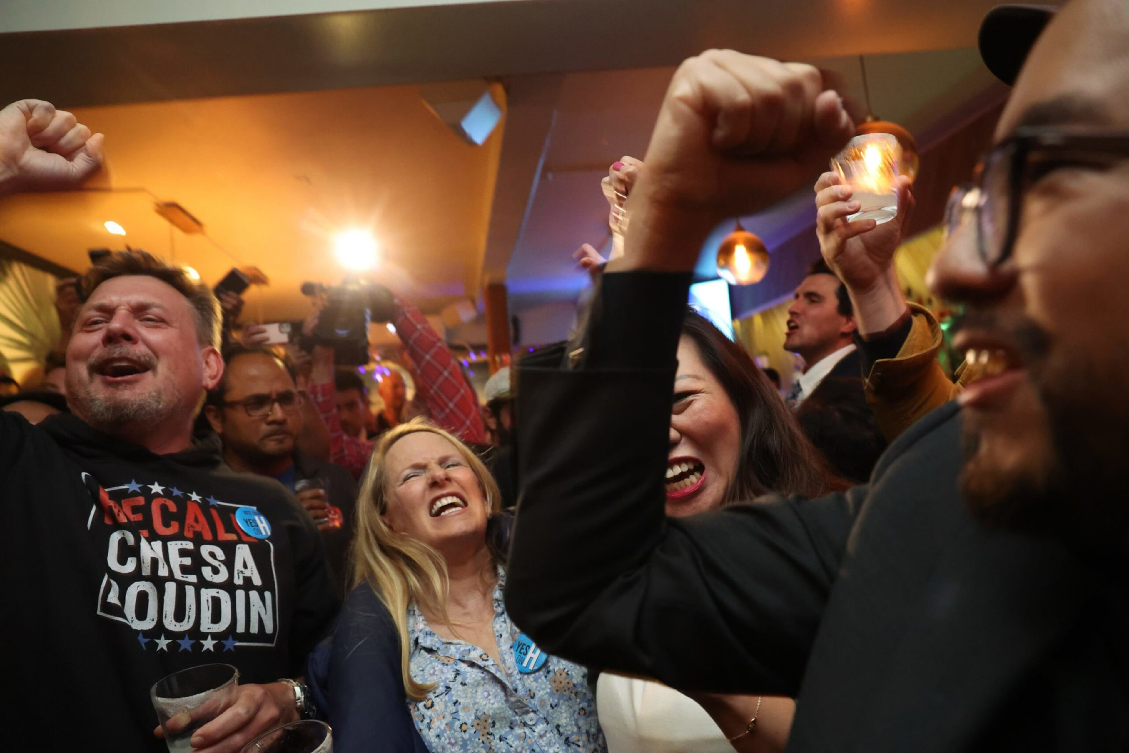 A crowd of diverse people cheer and raise their fists in celebration, with some holding drinks and candles, inside a warmly lit room.