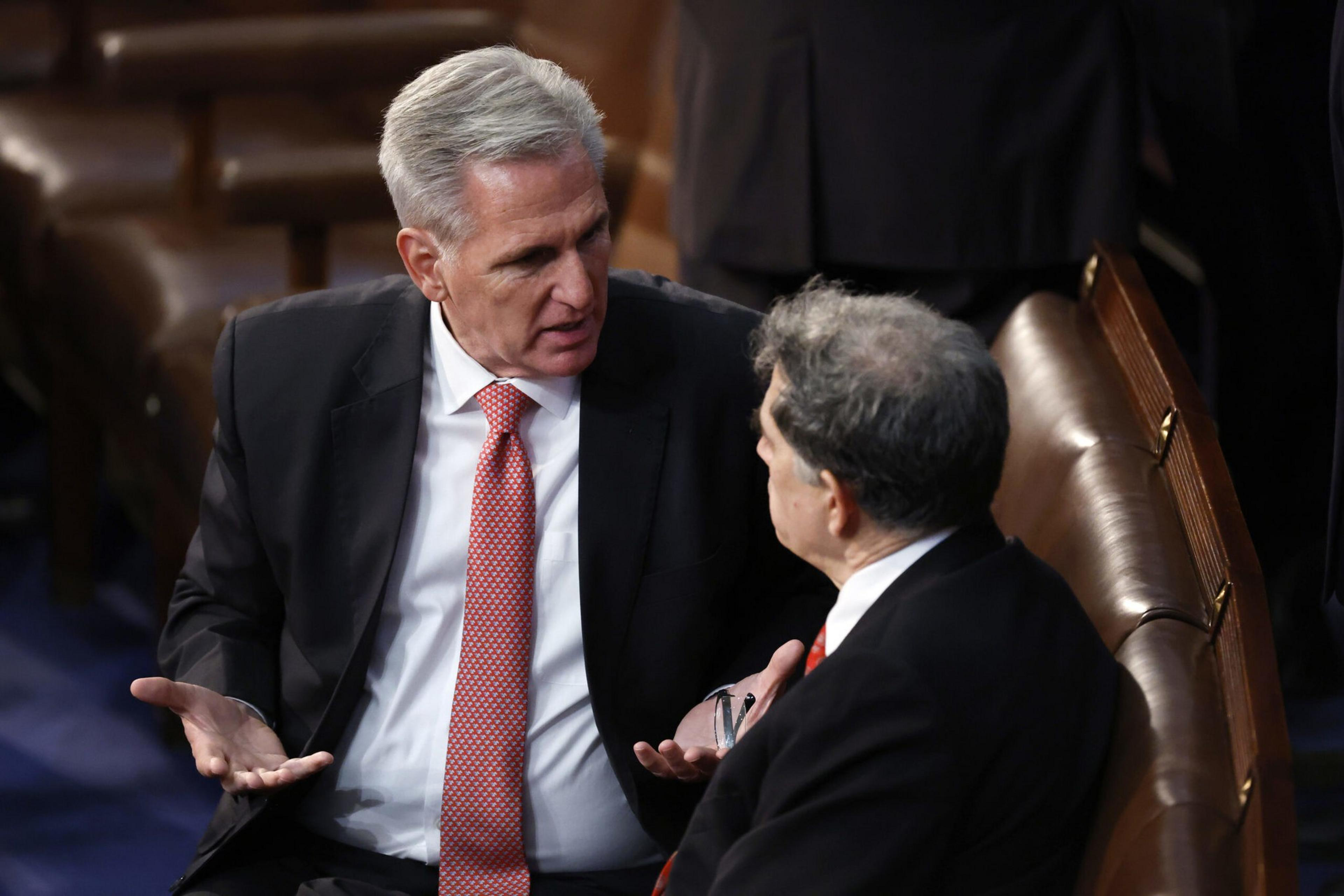 Two men in suits sit on leather chairs, engaged in a serious conversation, with one gesturing with his hands.
