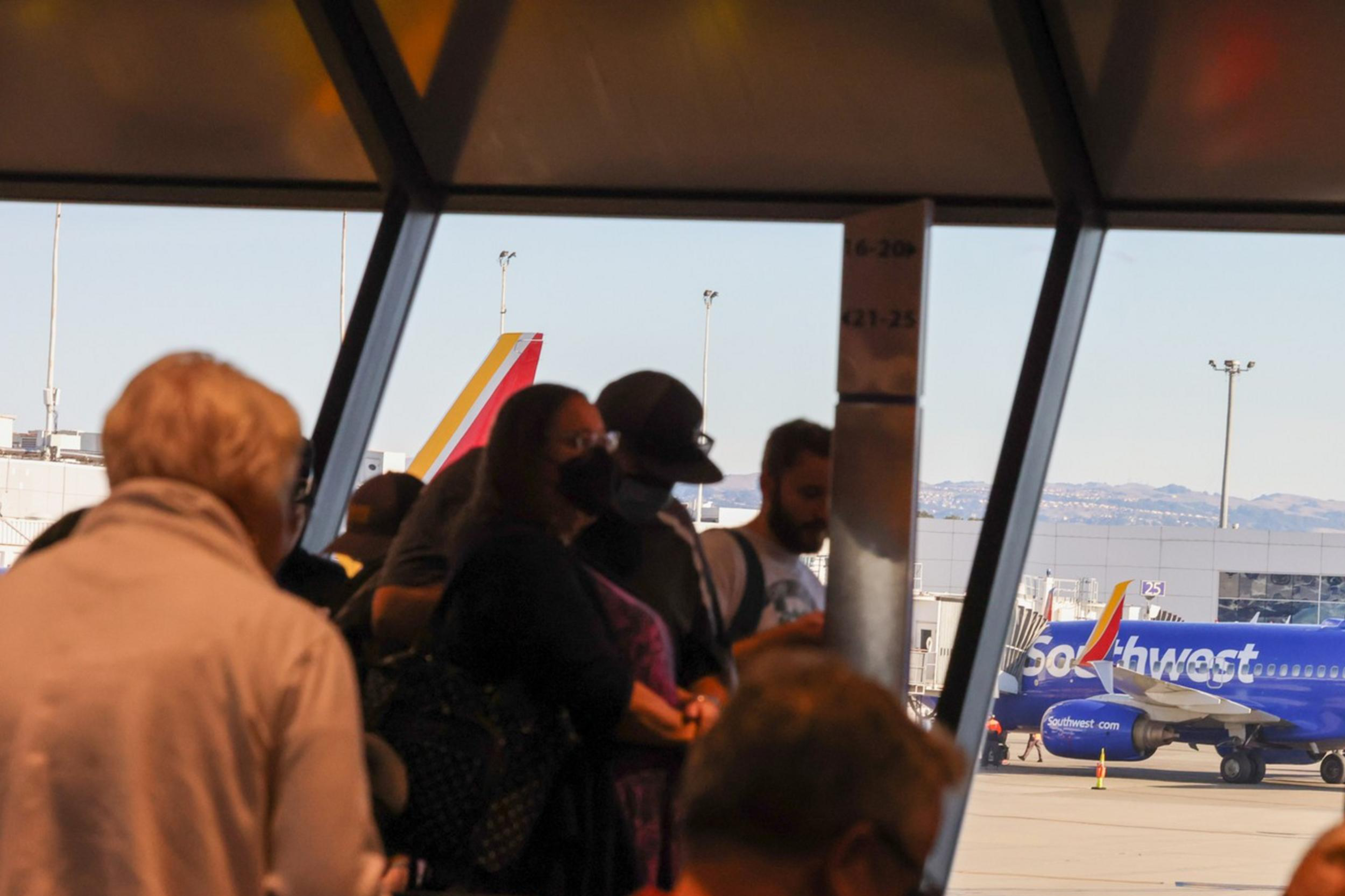 Passengers in a waiting area with a blue airplane outside the window and mountains in the backdrop.