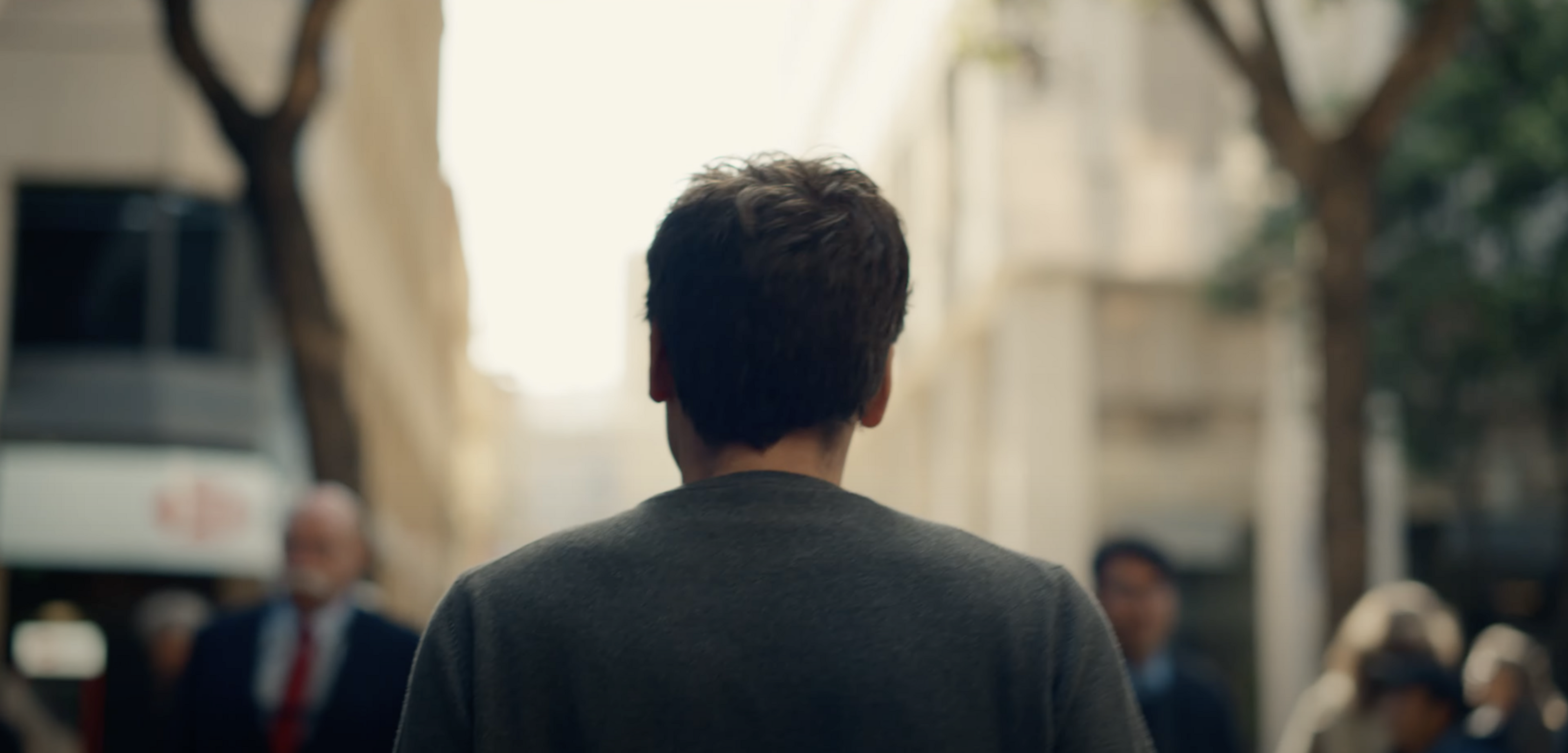 A man with short hair is walking down a blurred city street. He's wearing a gray shirt, and several people are around him, also out of focus.