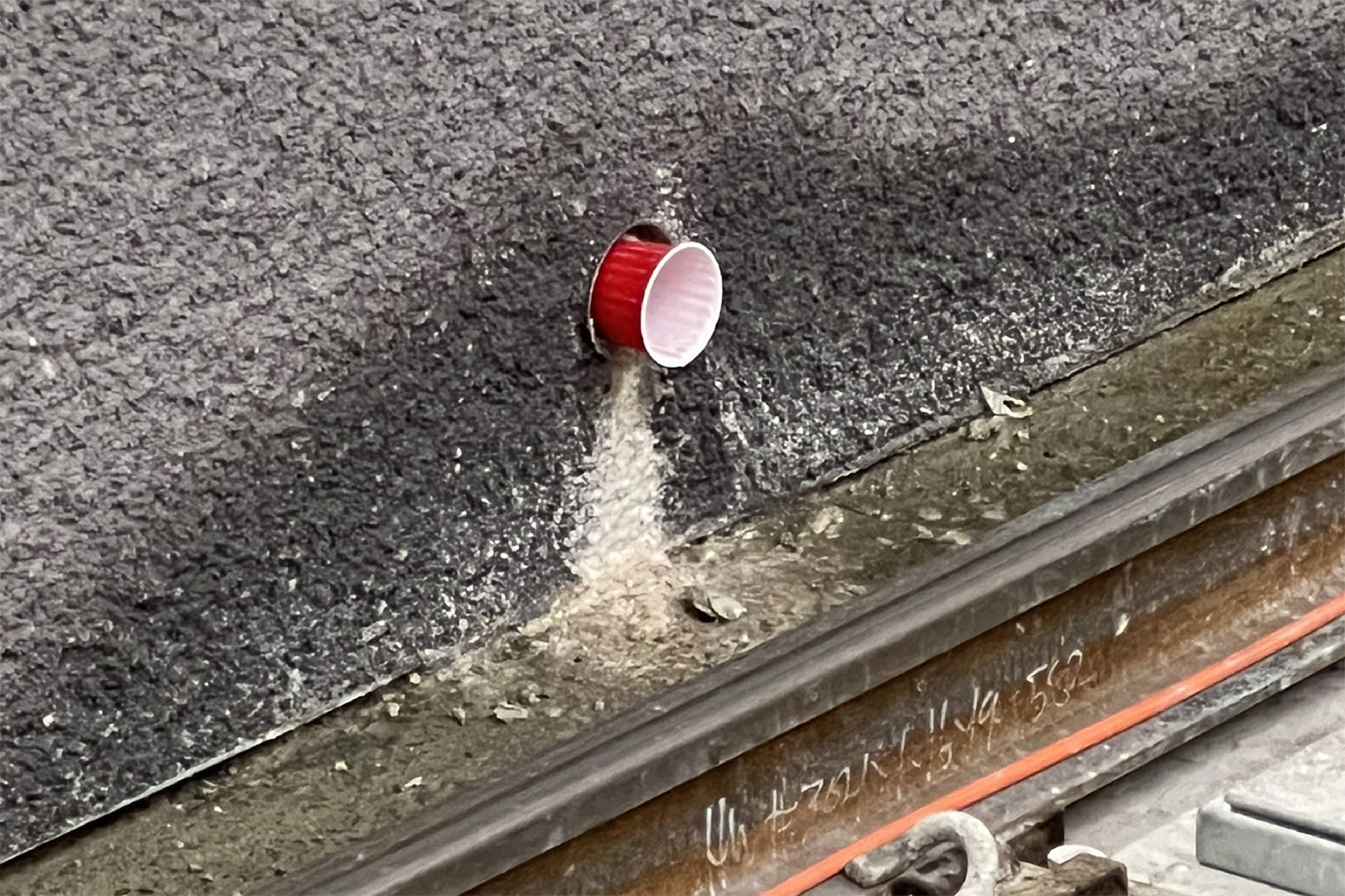 A red plastic cup is stuck on its side in a hole on a rough concrete wall above metal tracks. Dust and debris scatter below it.