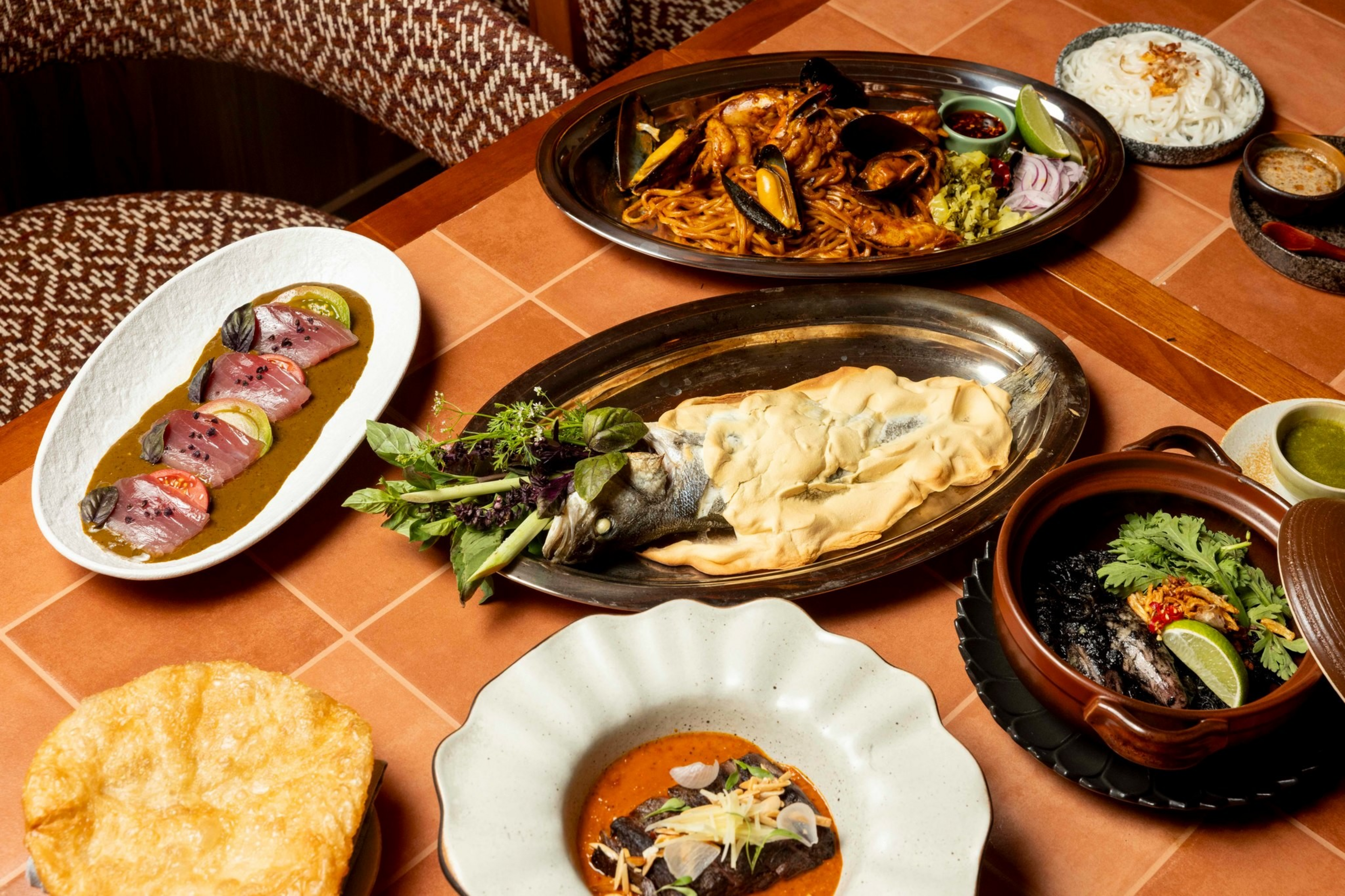 A table with assorted dishes including salt-crusted fish, seafood pasta, sliced tuna, rice, black stew, and a large crisp bread.