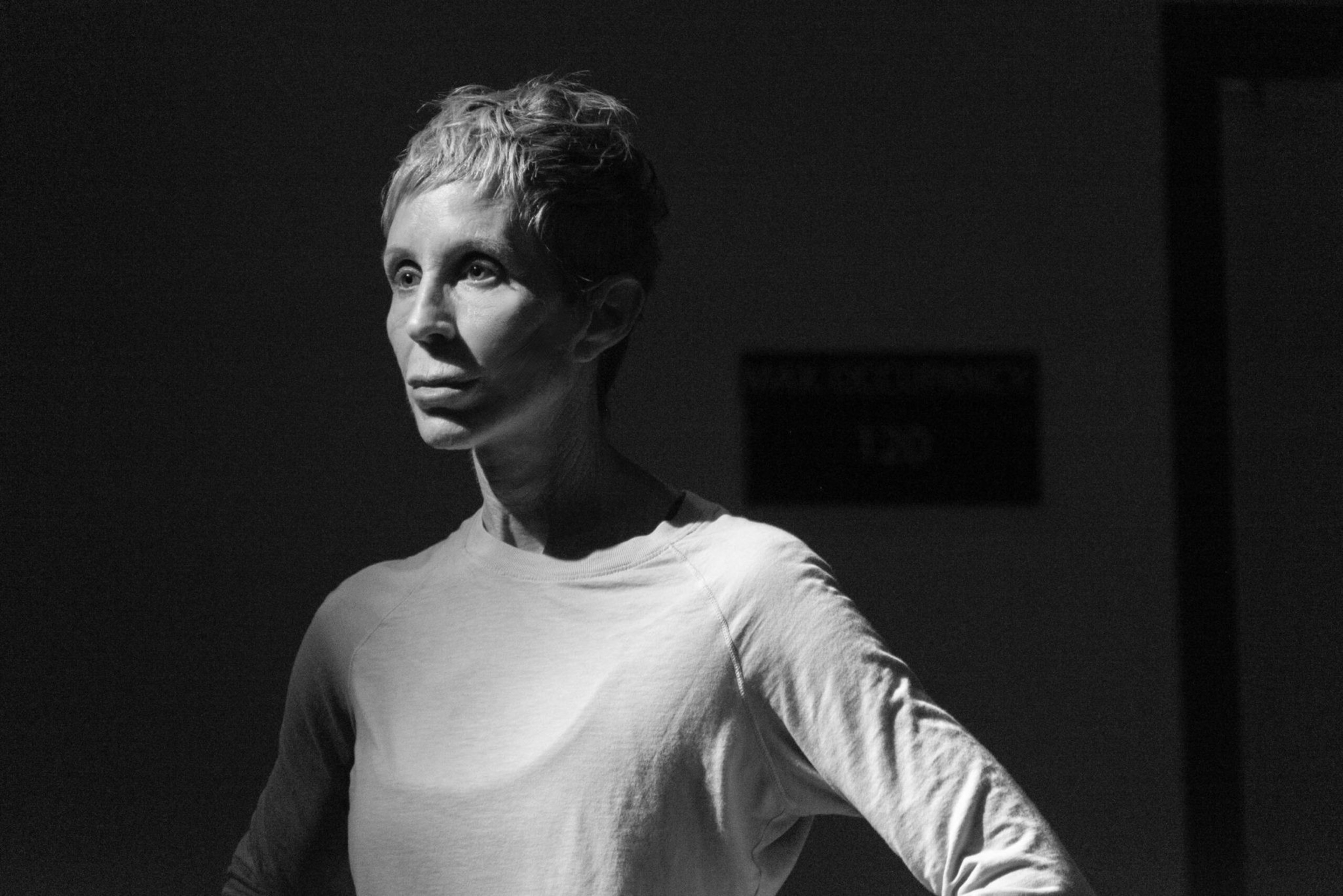A person with short hair wears a plain light shirt, standing with hands on hips, looking solemnly to the side in soft dramatic lighting.