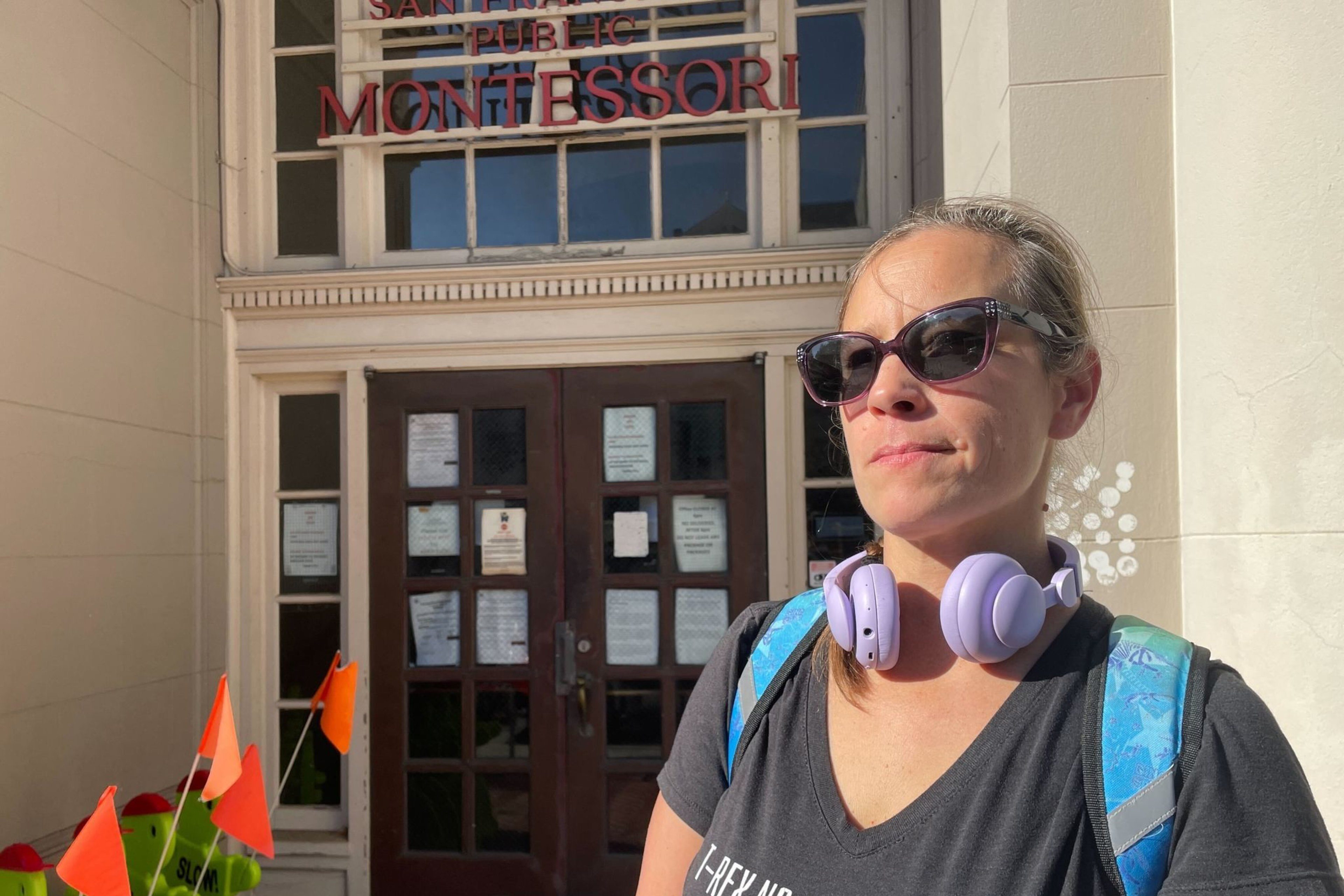 A woman wearing sunglasses, headphones, and a T-shirt stands in front of a building with a sign that reads "Montessori." There are small orange flags nearby.