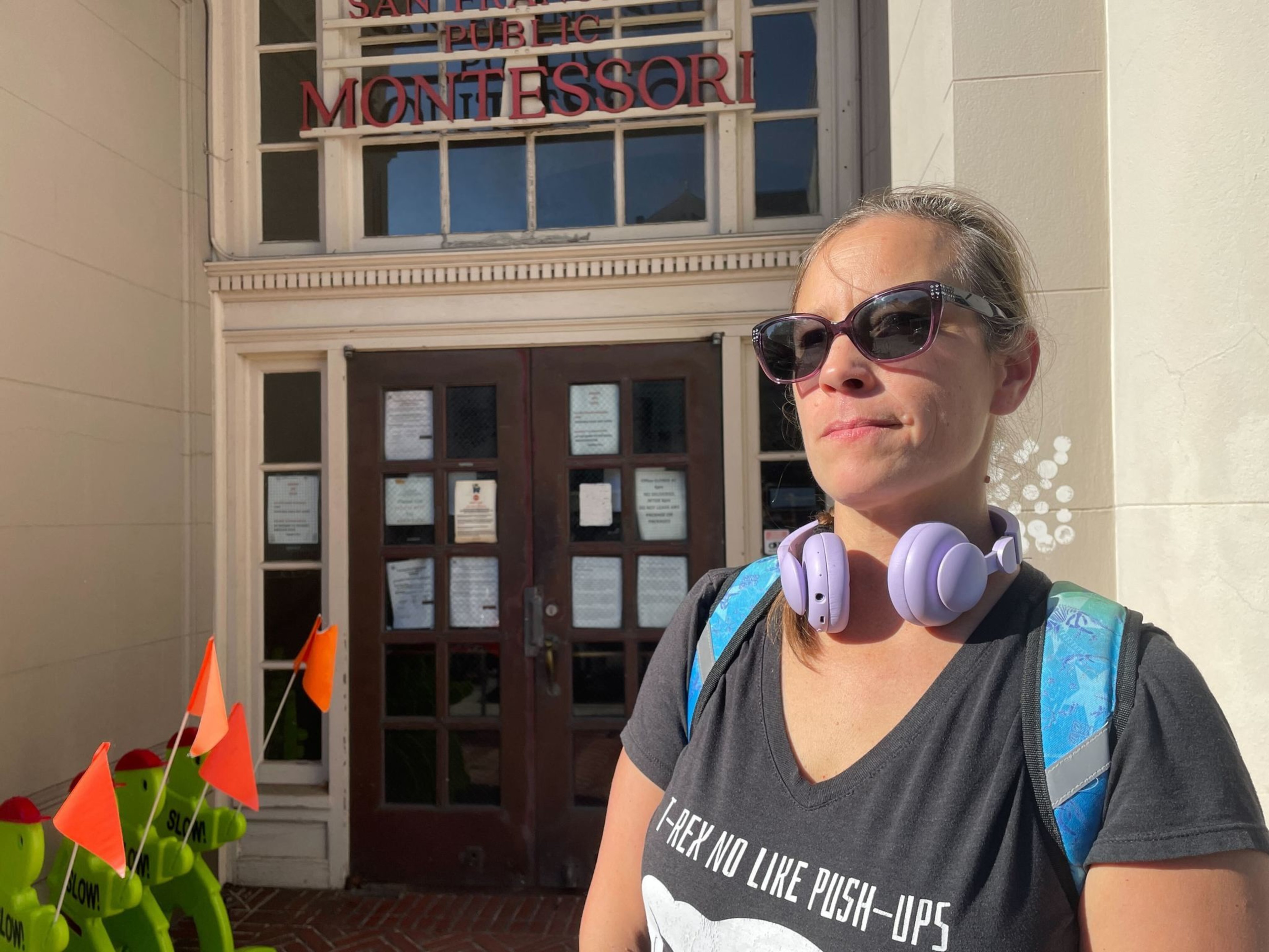 A woman wearing sunglasses, headphones, and a T-shirt stands in front of a building with a sign that reads "Montessori." There are small orange flags nearby.