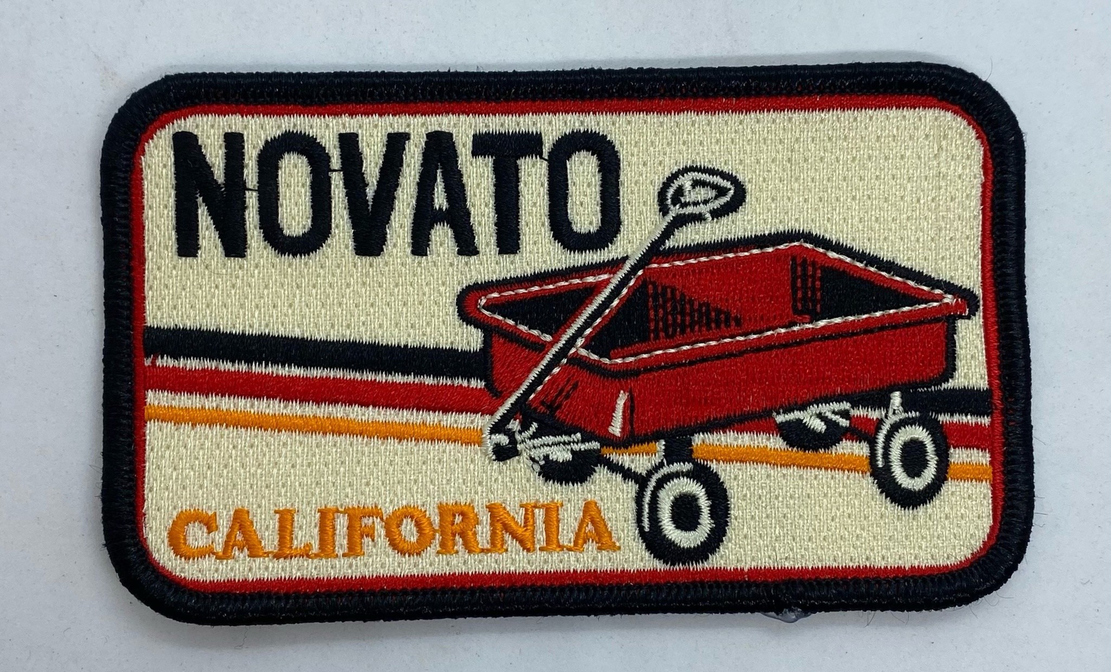A rectangular patch with a red wagon illustration, the word "NOVATO" in black at the top, and "CALIFORNIA" in orange at the bottom.