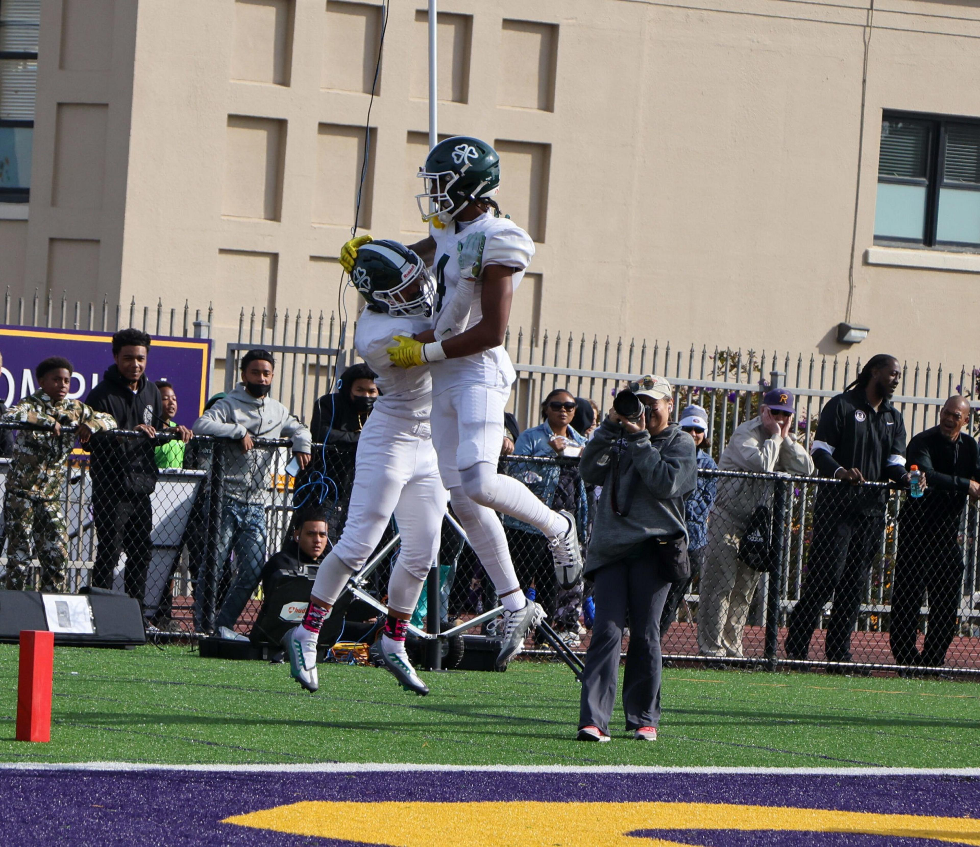 Two football players in white uniforms and black helmets jump and celebrate in the end zone while a photographer captures the moment nearby.