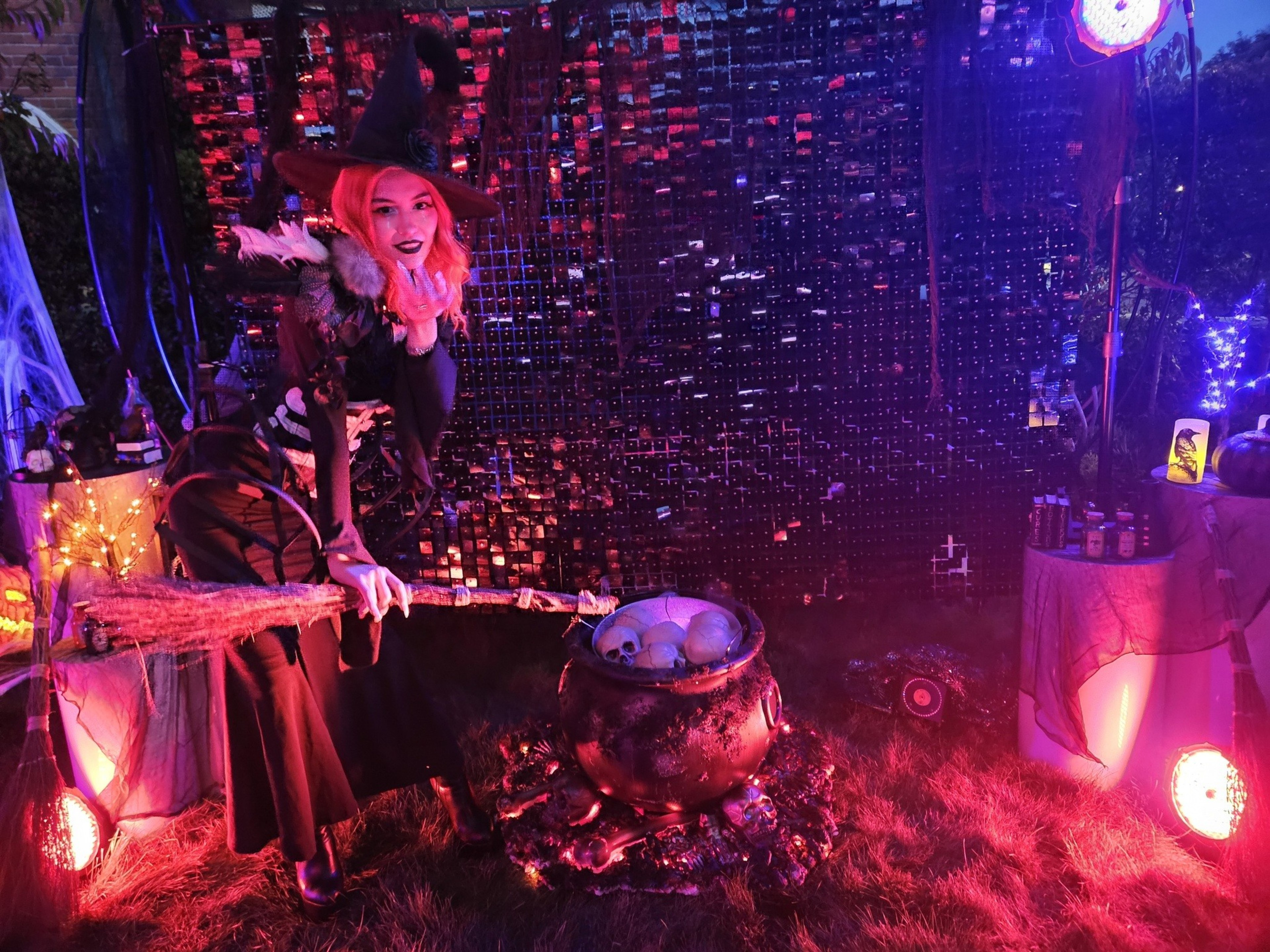 A woman dressed as a witch is posing with a broom in a Halloween scene. She stands next to a cauldron with skulls, surrounded by spooky decorations and eerie lighting.