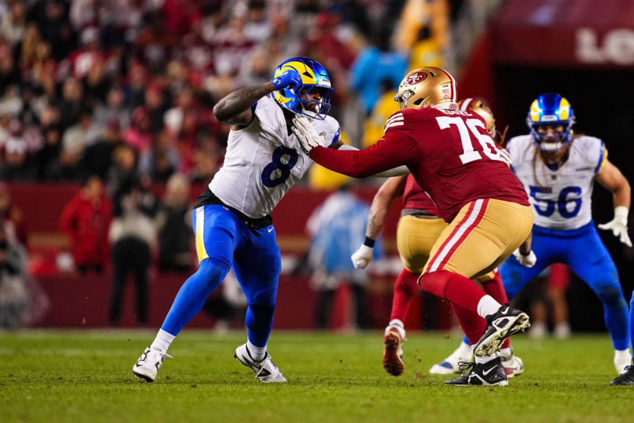 A football player in a white and blue uniform blocks another player wearing red and gold. The action is intense, surrounded by a blurred crowd in the background.