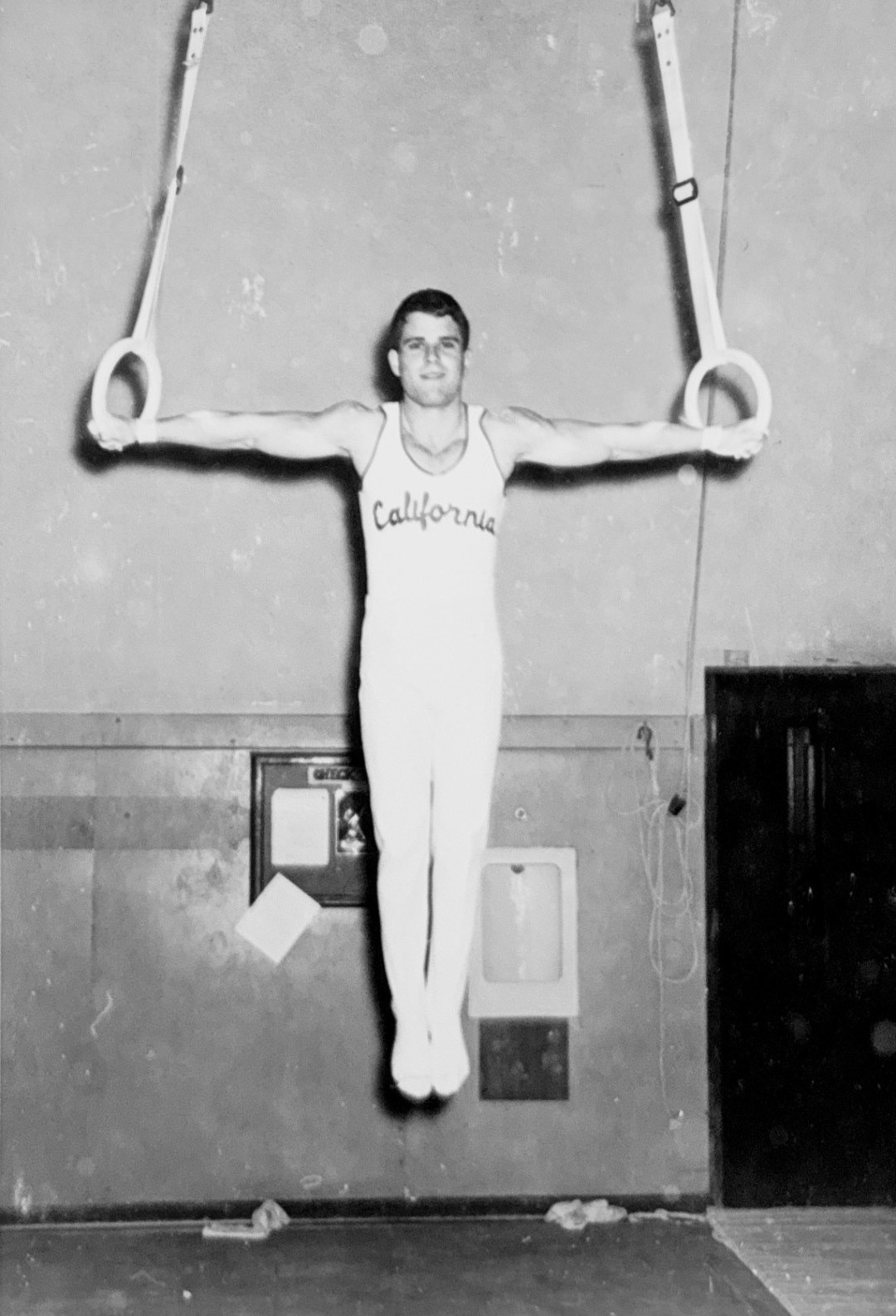A male gymnast in a “California” singlet holds a still iron cross pose on gymnastic rings in an indoor gym.