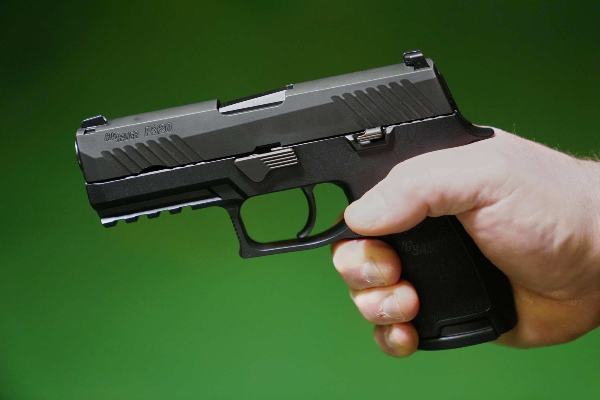 A person holds a black Sig Sauer P320 handgun against a green background, gripping it firmly with their right hand.