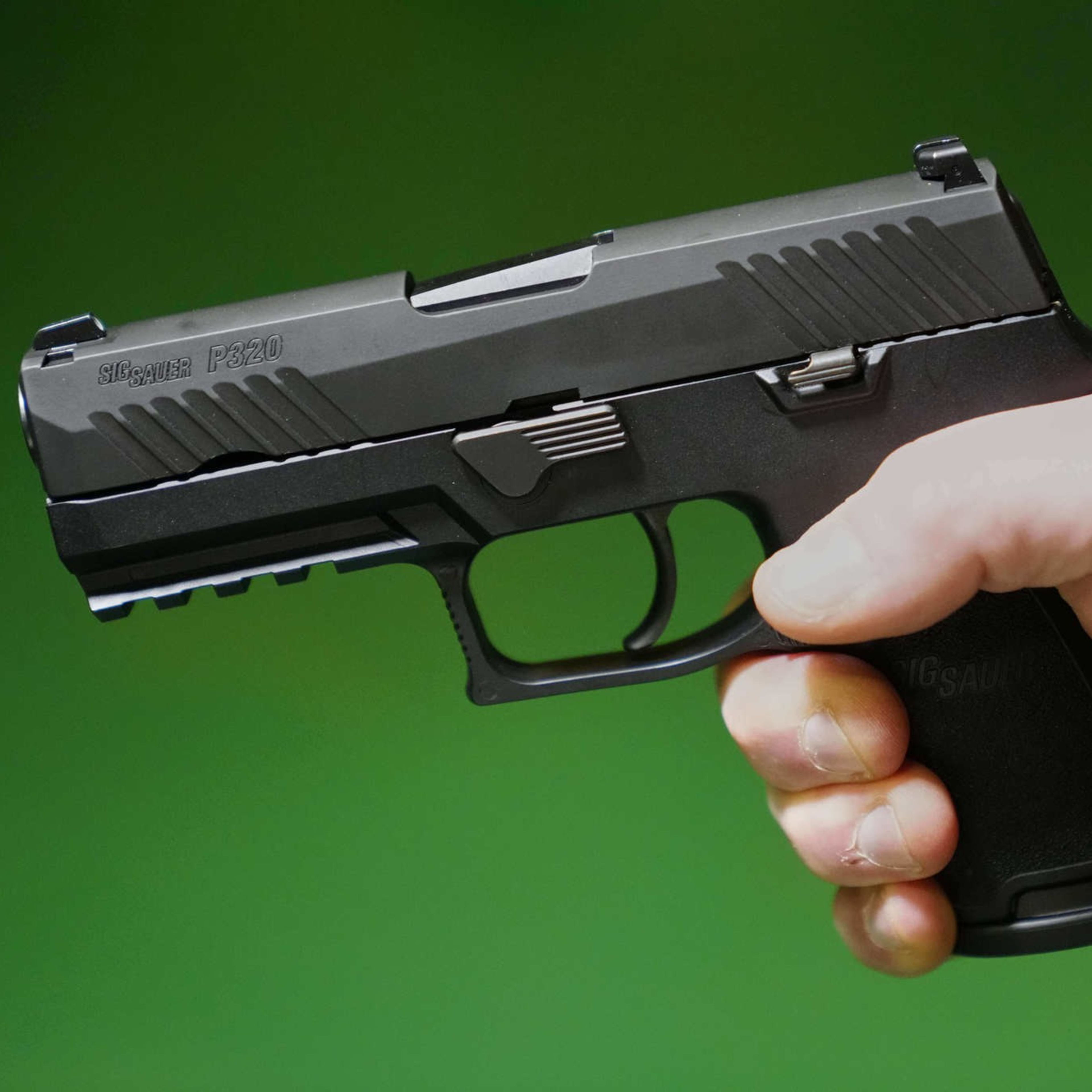 A person holds a black Sig Sauer P320 handgun against a green background, gripping it firmly with their right hand.