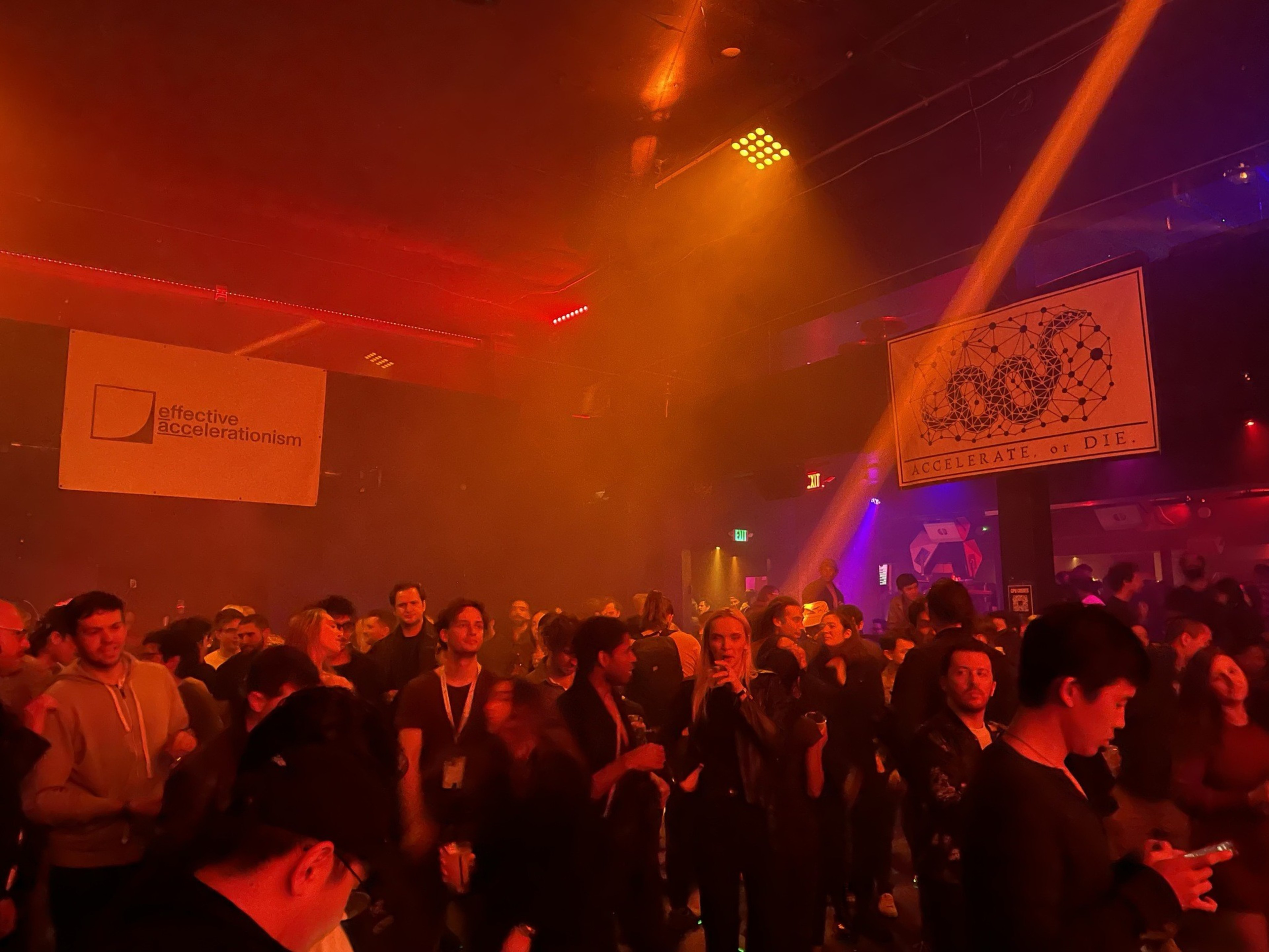 Hundreds of people stand on a nightclub dance floor bathed in red light with white posters reading &quot;effective accelerationism&quot; and &quot;accelerate or die&quot; hanging above them.