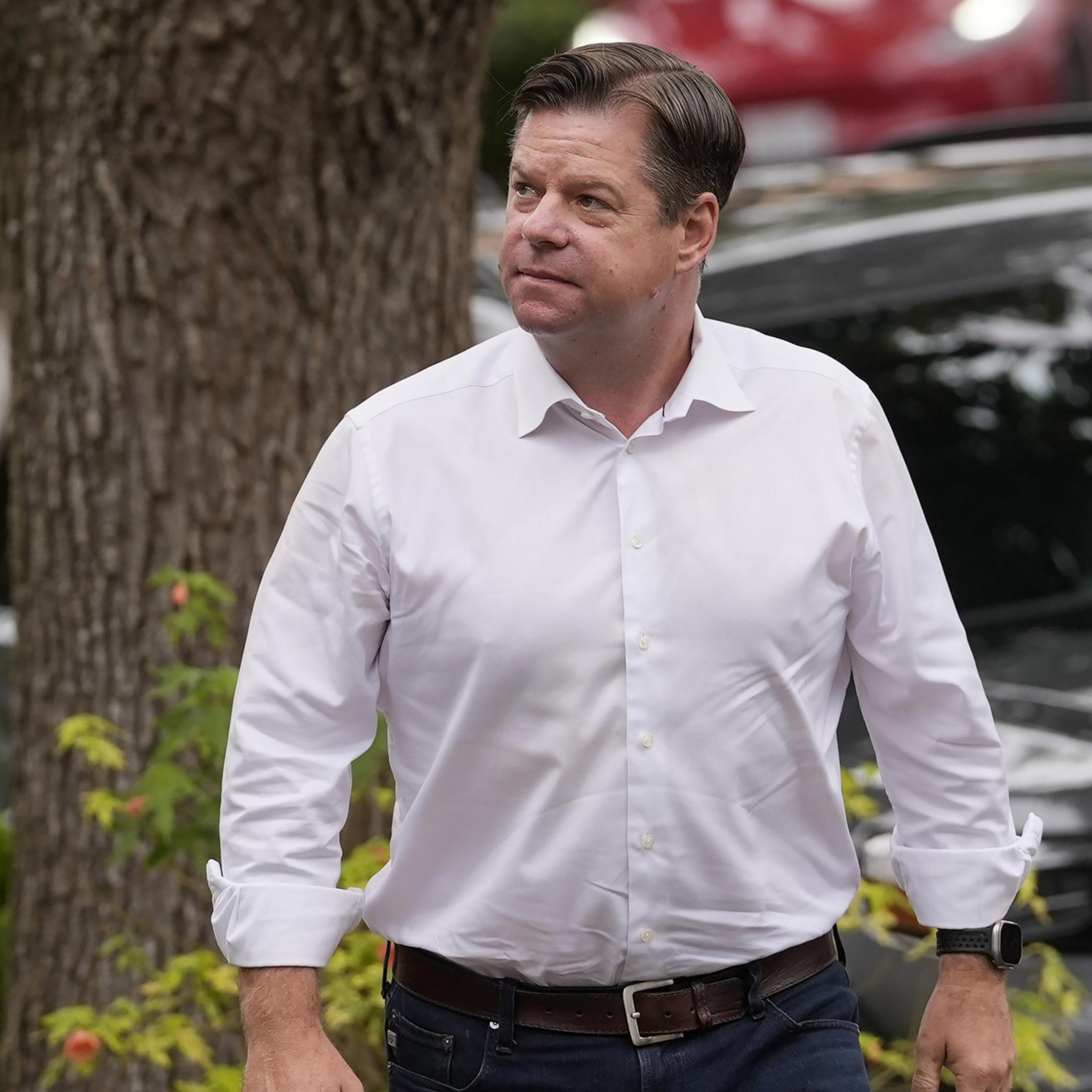 A man in a white shirt and jeans walks outside near a parked black car and greenery. He's looking slightly upwards, with trees and other cars in the background.