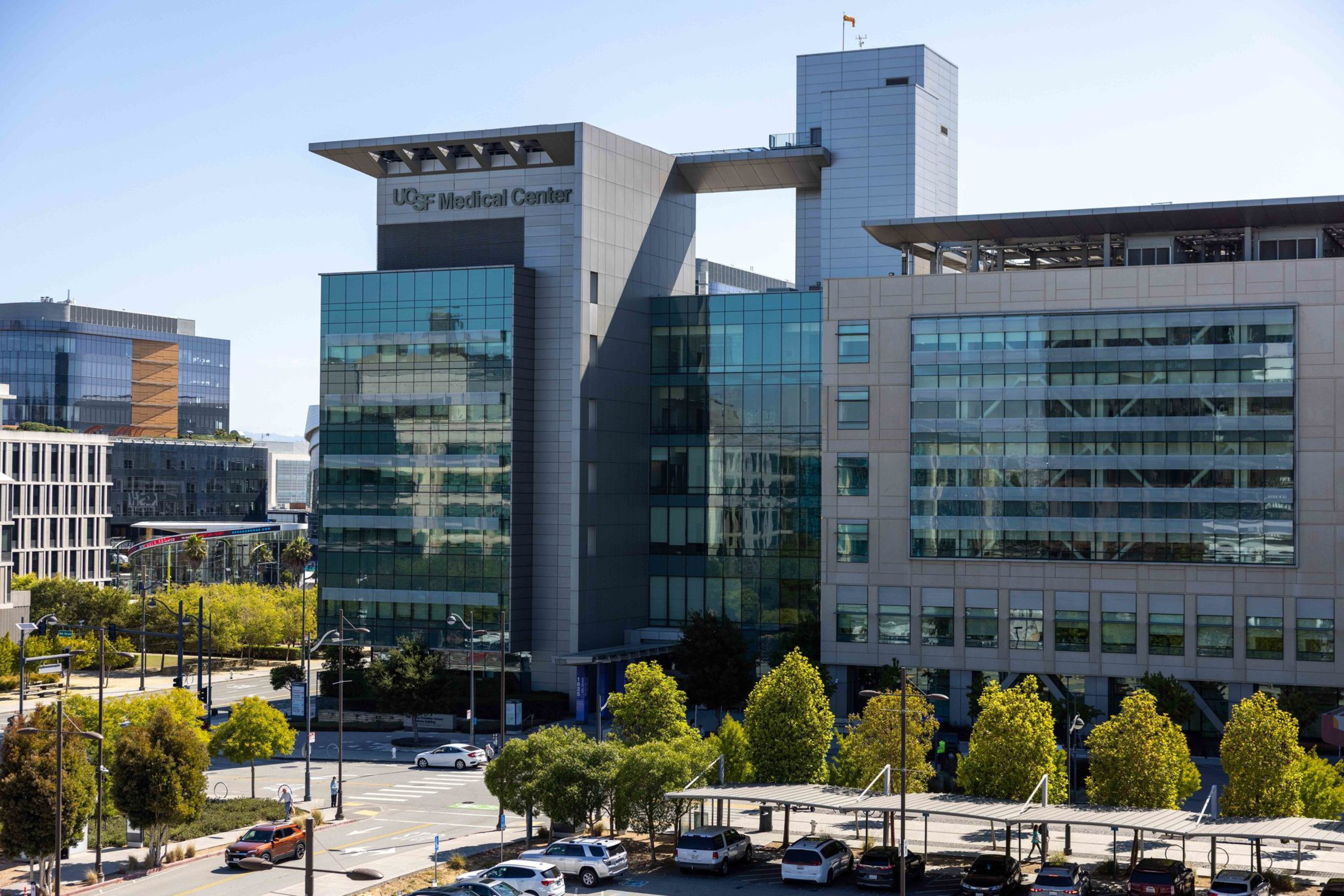 A modern glass and concrete building labeled “UCSF Medical Center” with trees, parked cars, and streets in the foreground under a clear sky.