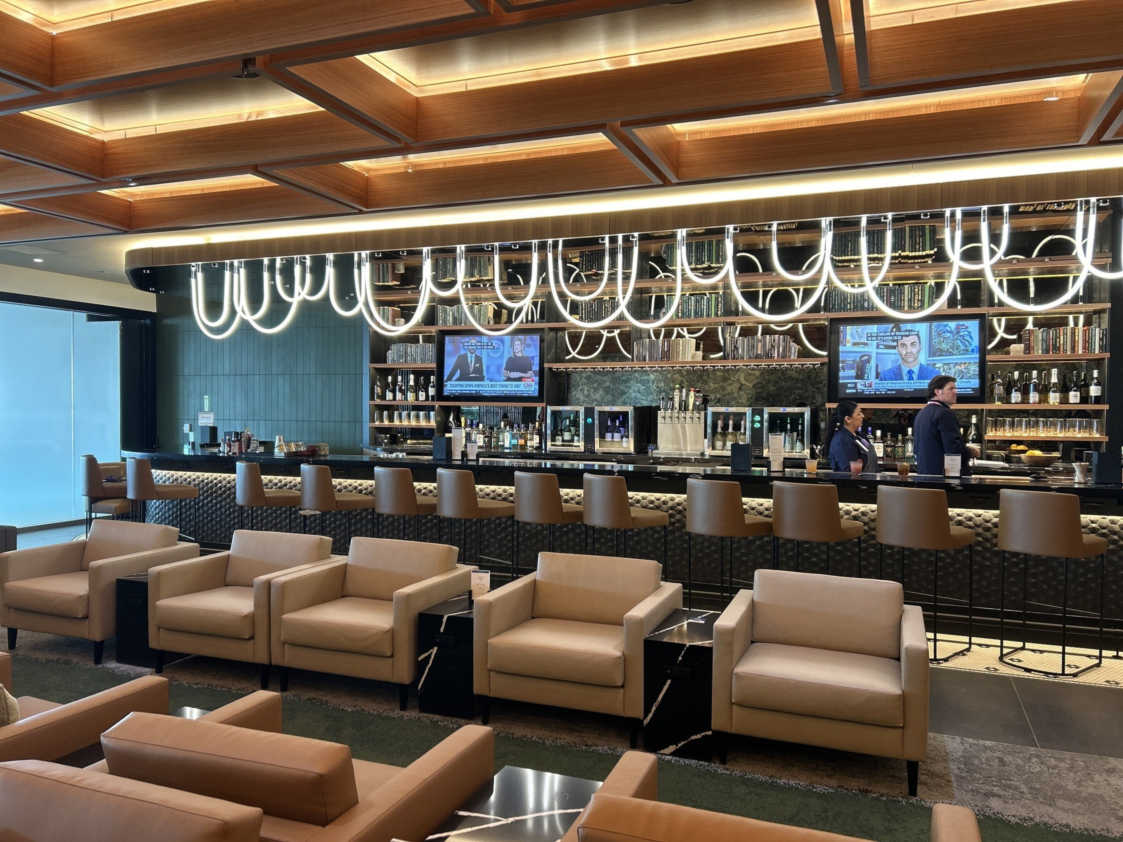 The image shows a modern bar with a stylish, illuminated ceiling, tan leather chairs, and high stools at the bar, where two people are working. Two TVs display news.