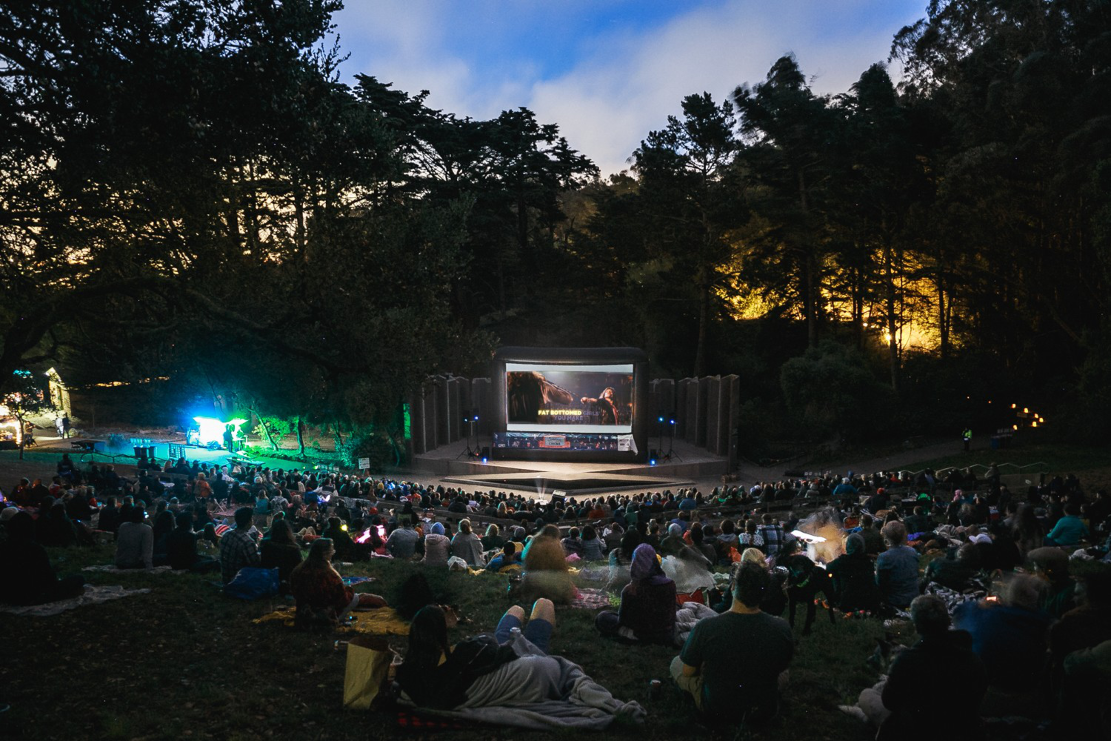 A large crowd sits on grassy hill watching an outdoor movie screening at dusk, surrounded by tall trees and illuminated stage lights.