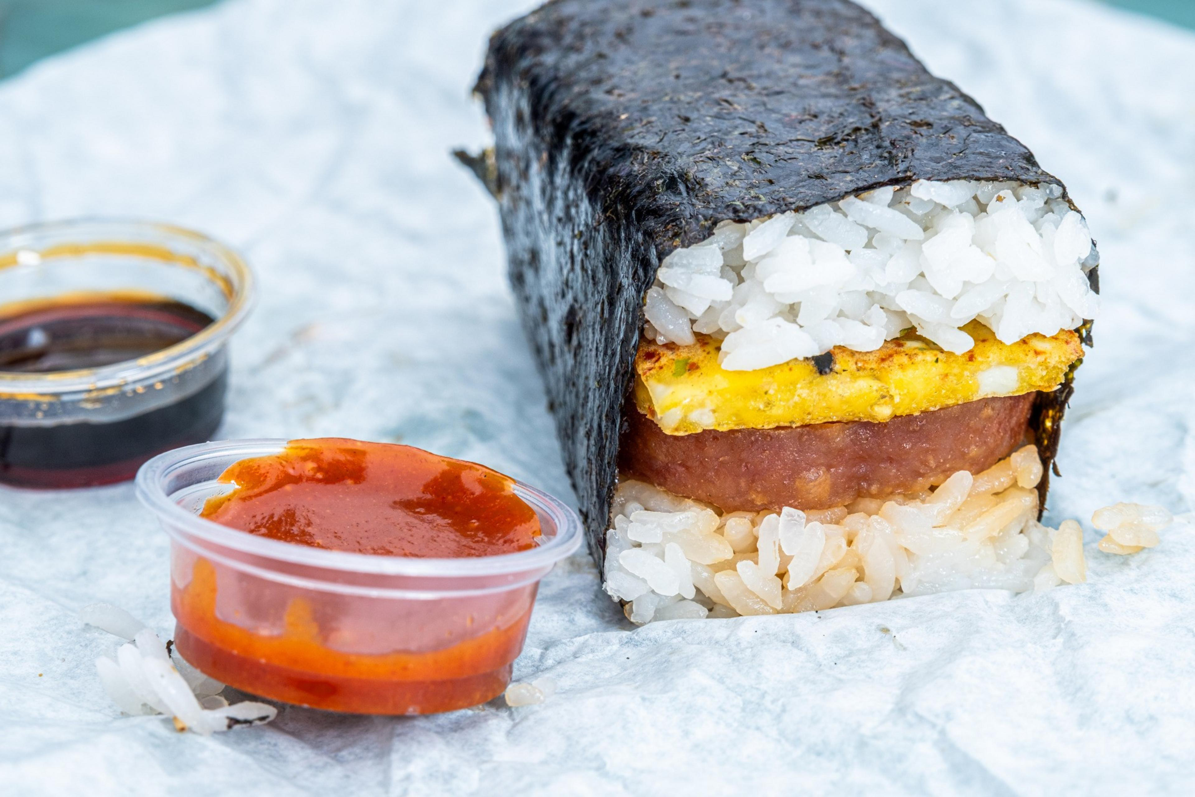 The image shows a musubi, featuring rice, egg, and a slice of meat wrapped in seaweed, placed on a paper. Two sauce containers are nearby.