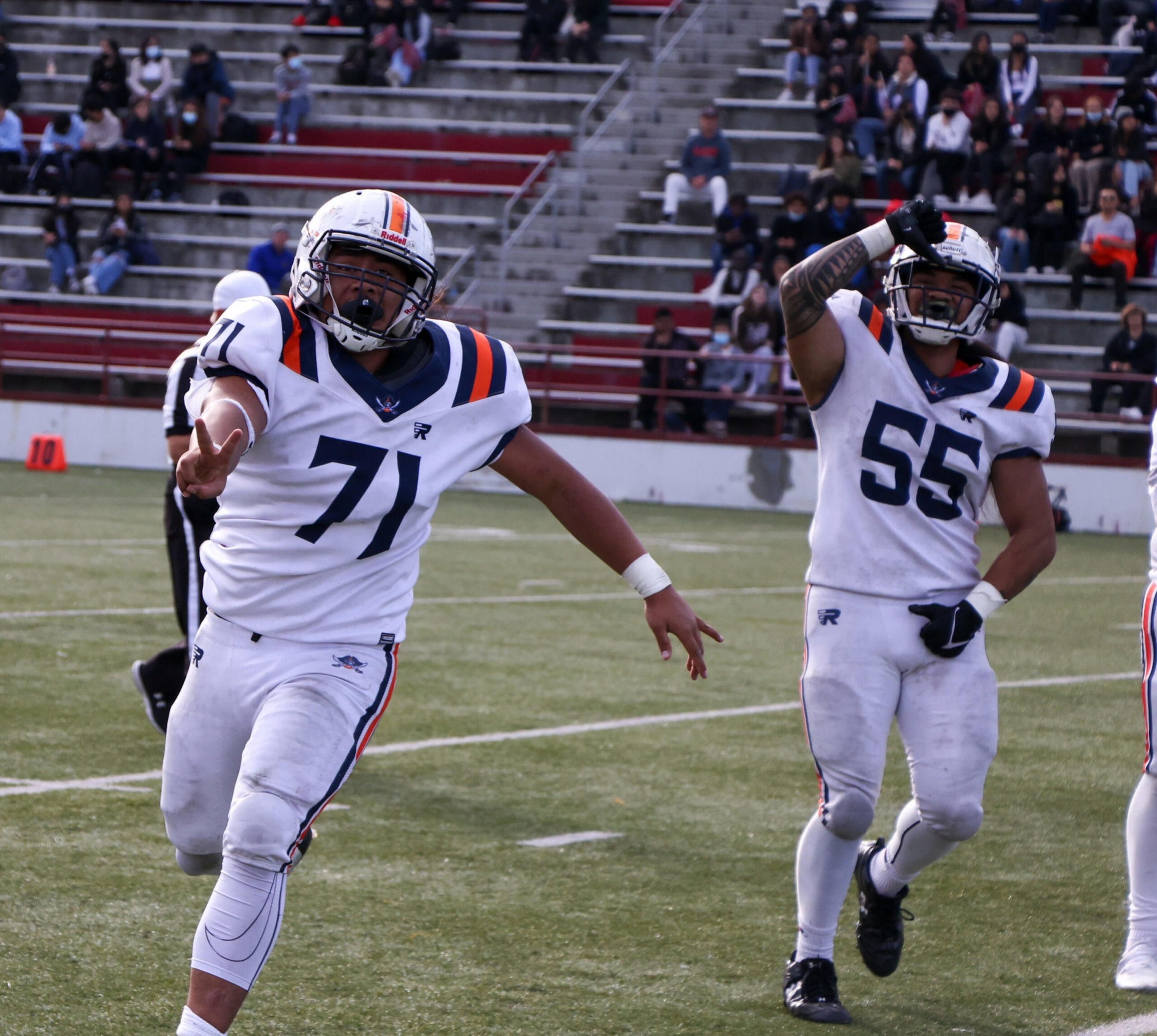 Two American football players in white and navy uniforms celebrate on the field with fans in the stands behind them.