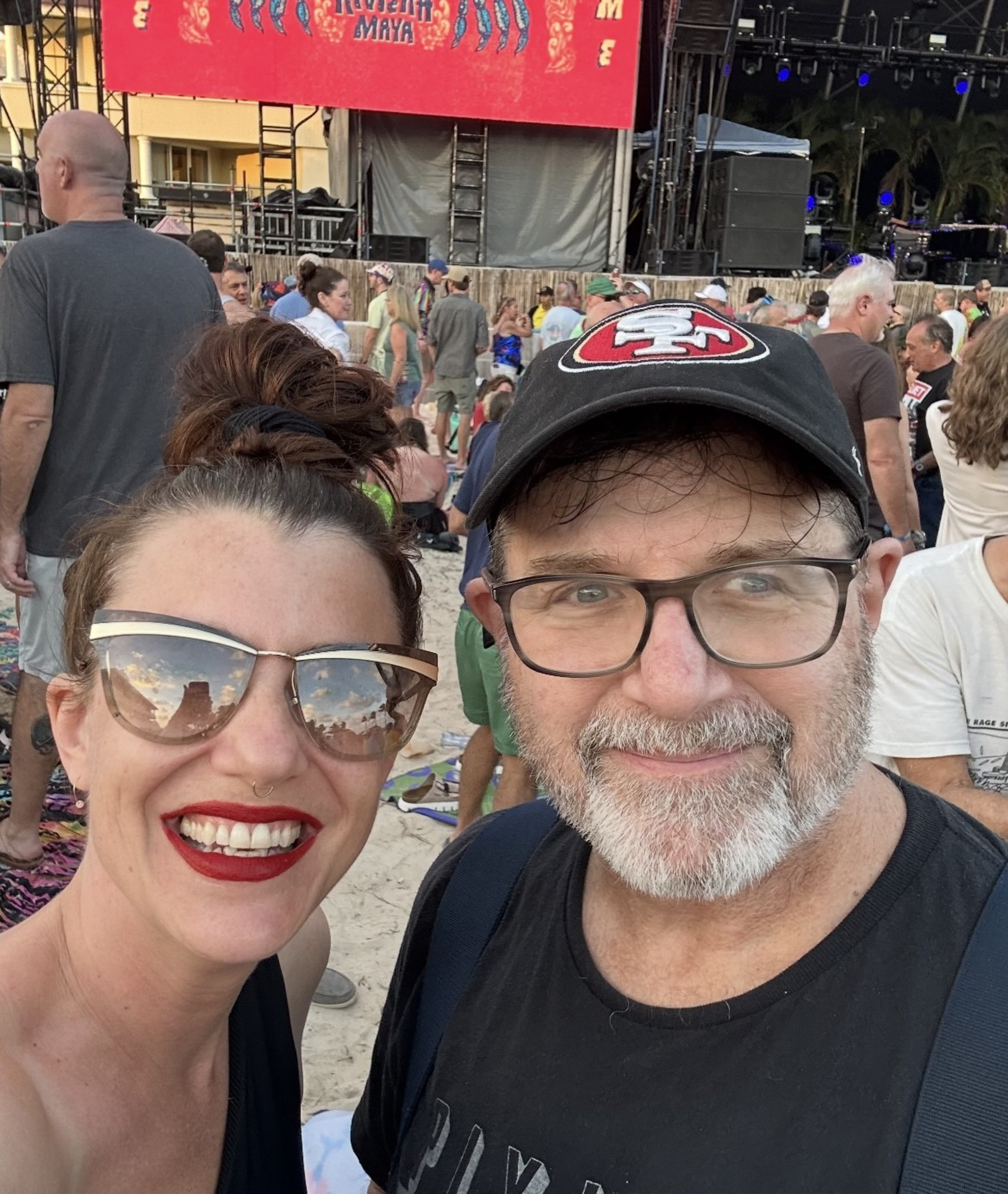 A smiling woman and man are at an outdoor event. She wears sunglasses and red lipstick; he has glasses and a hat. There are many people and a stage behind them.