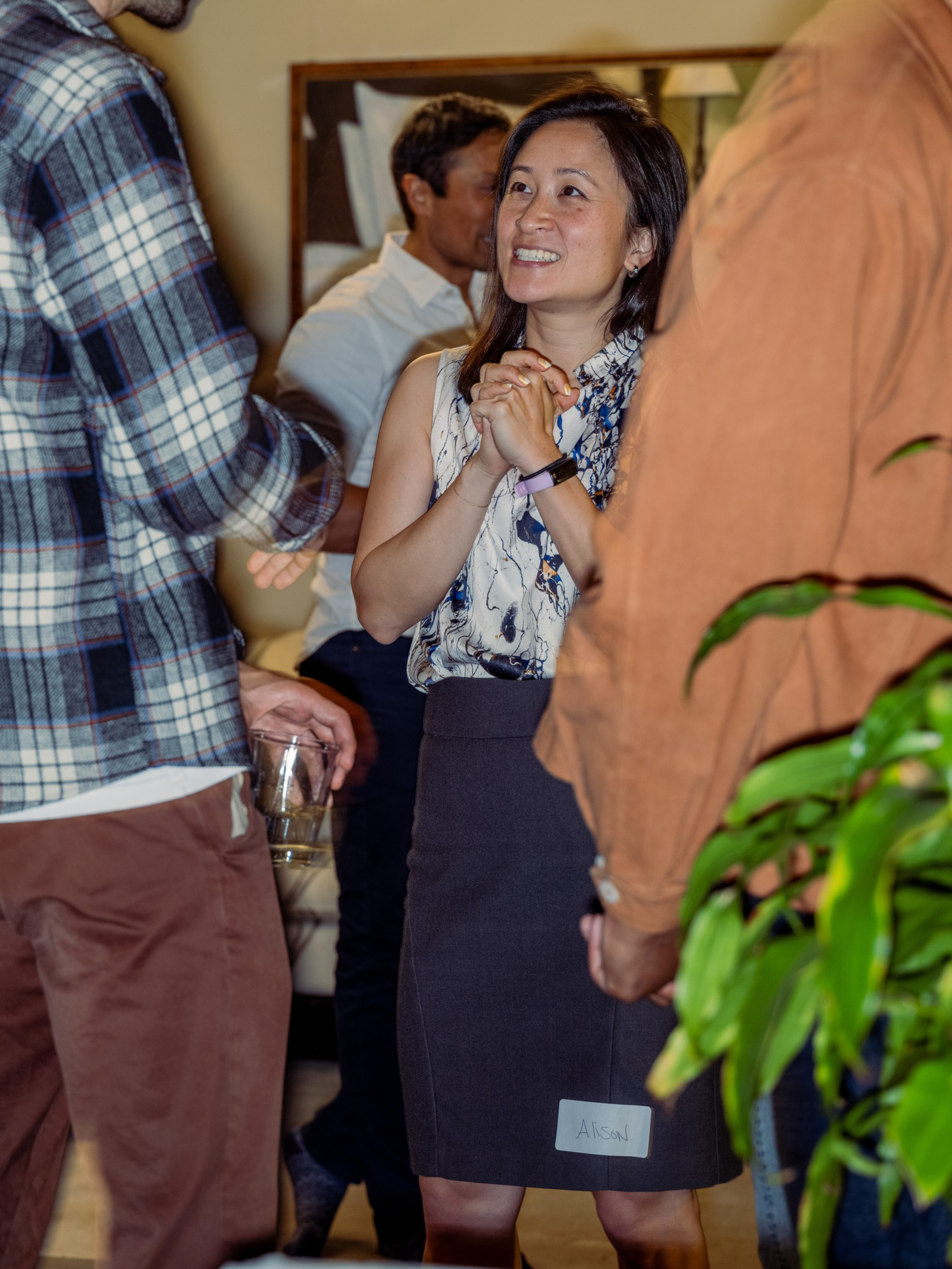 A smiling woman with clasped hands talks to two men, one holding a glass, at an indoor social gathering with plants and framed artwork.