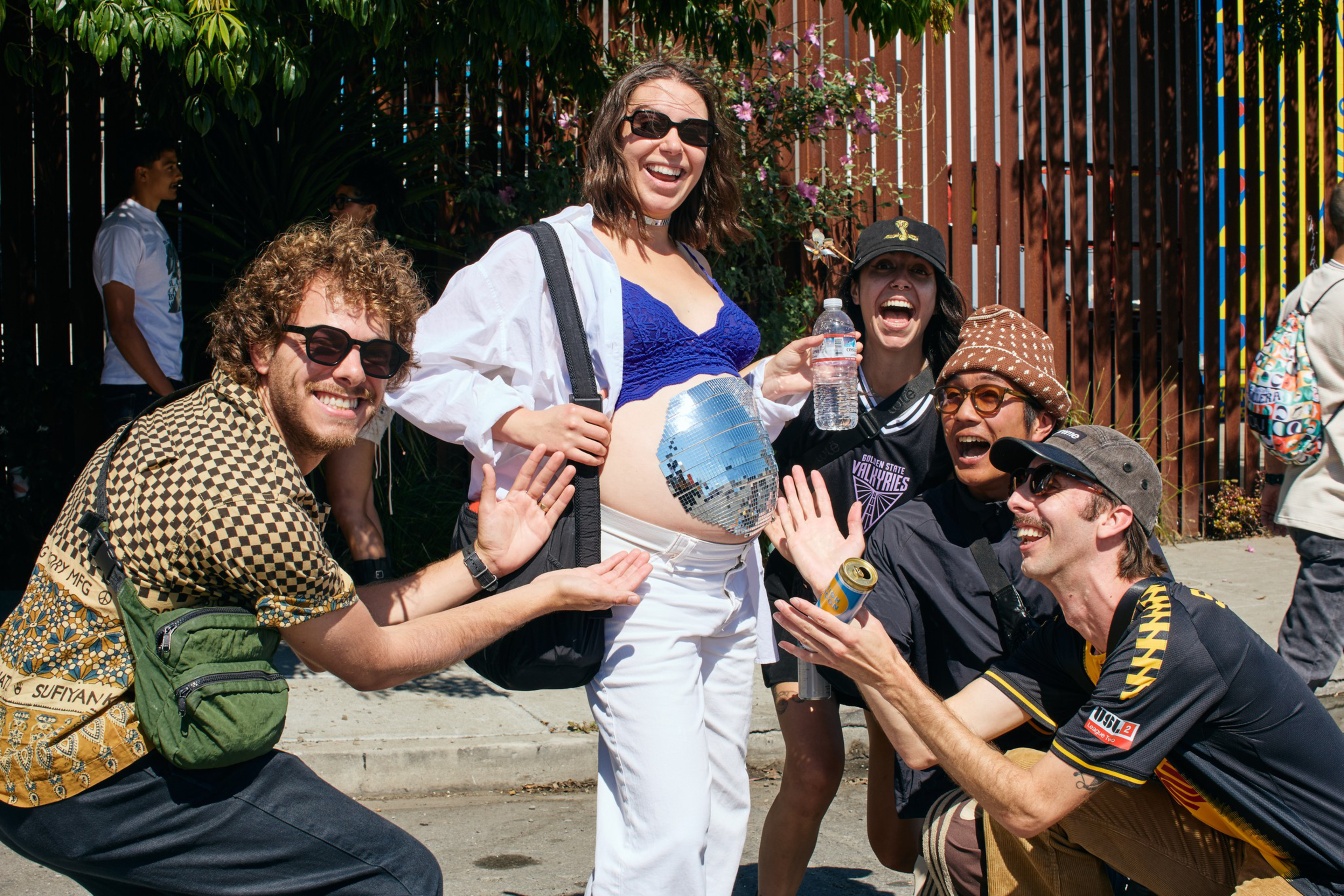 Five friends smile and pose outdoors, one wearing sunglasses and white pants with a shiny disco ball attached to their exposed belly.