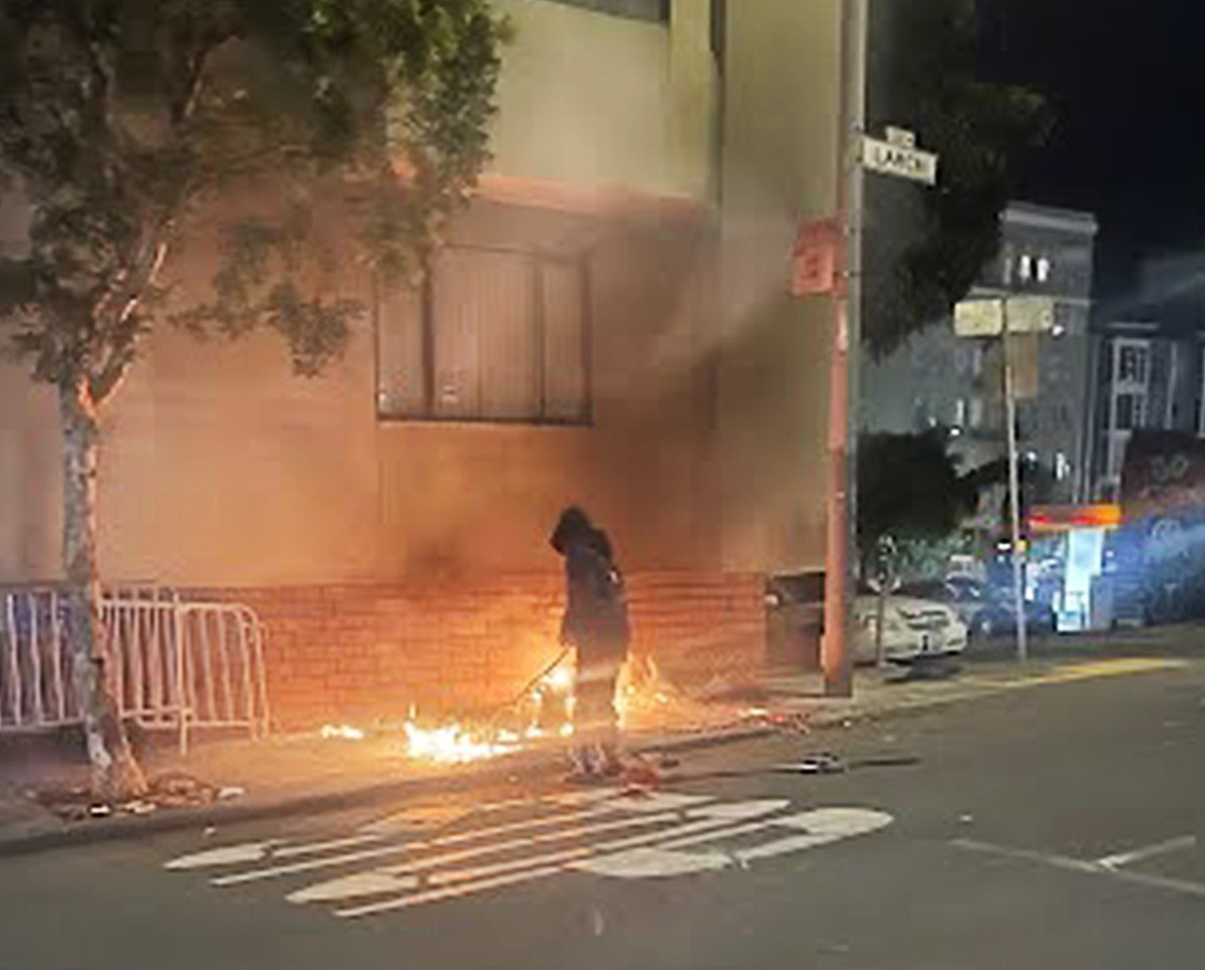 A person stands near small fires on a city sidewalk at night beside a building under a tree, with street signs and cars visible.