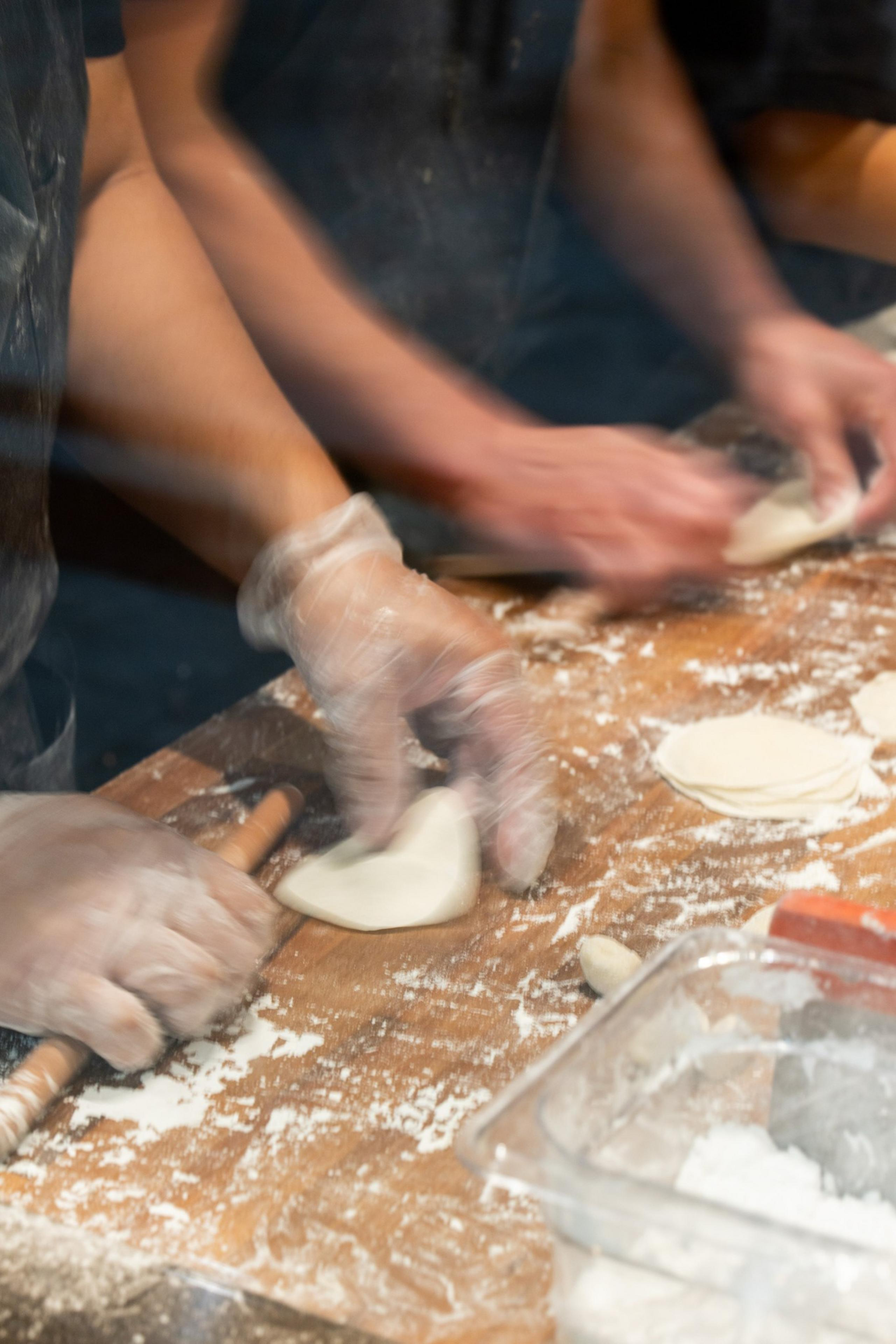 People are making dough on a floured surface, using their hands and rolling pins. They're wearing gloves, and there are several round dough pieces.