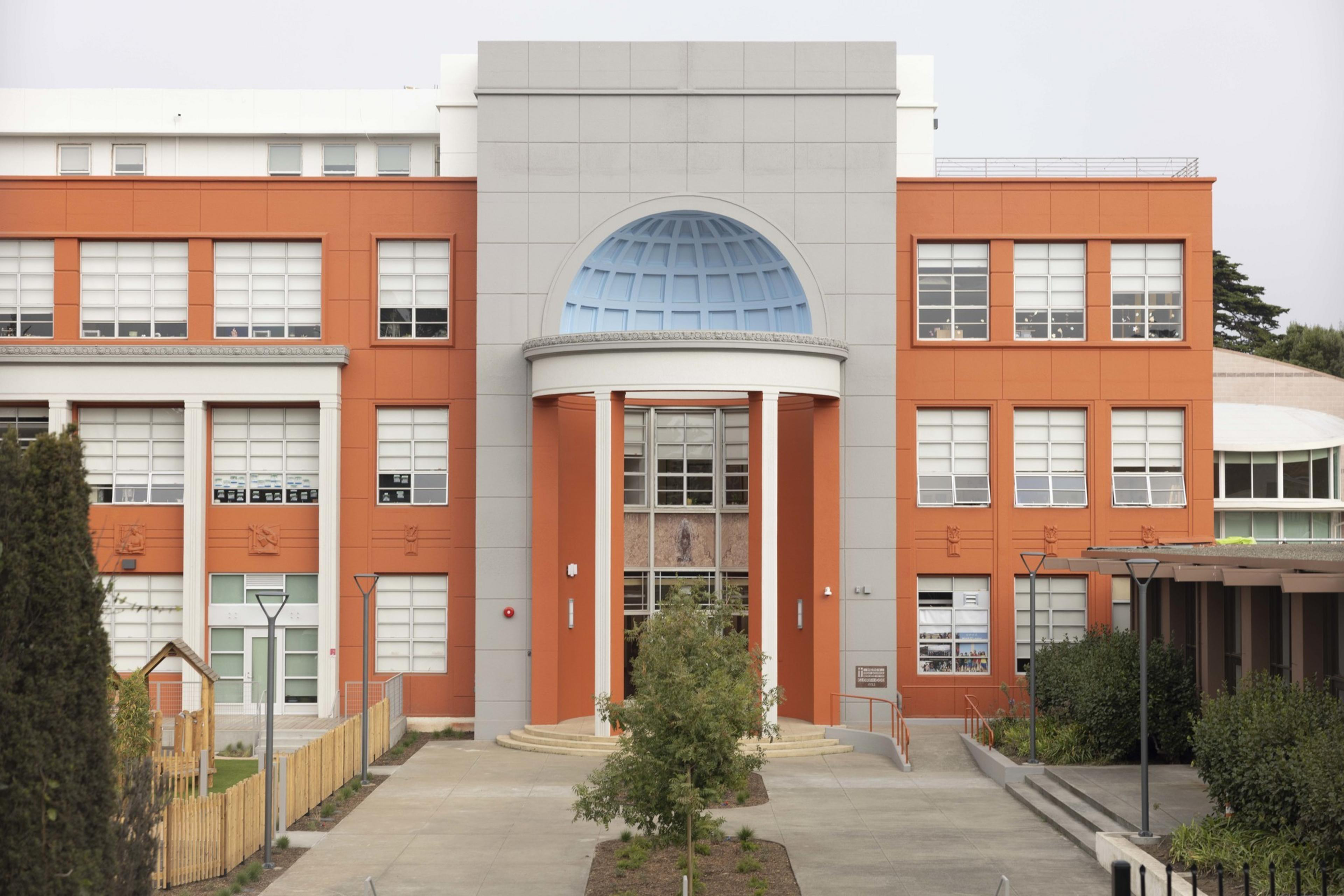 The image shows a modern building with an orange and gray facade, featuring large windows and a prominent arched entrance, surrounded by greenery.