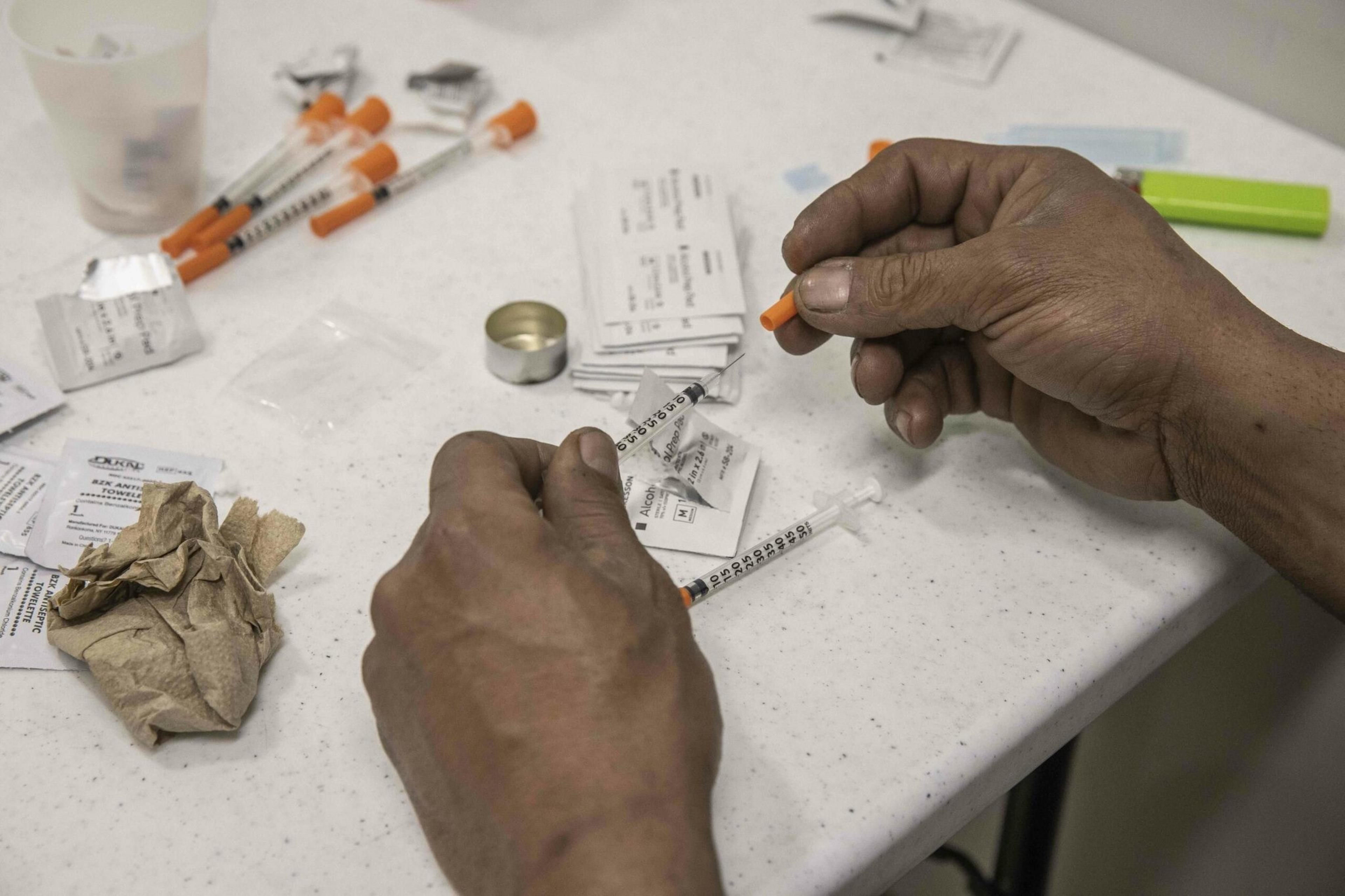 A pair of hands handling syringes with orange caps rest on a table scattered with medical packaging, a crumpled tissue, and a small metal cap.