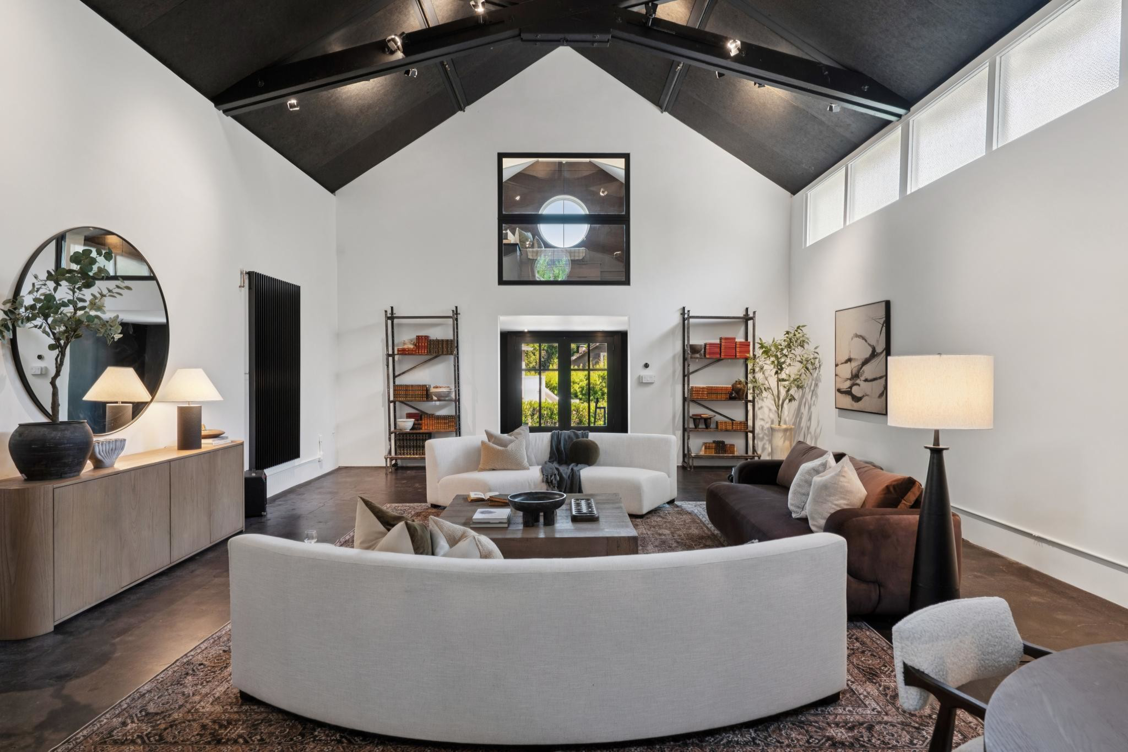A spacious living room with dark vaulted ceilings, white and brown sofas, tall bookshelves, a wooden coffee table, and soft lighting from lamps and windows.