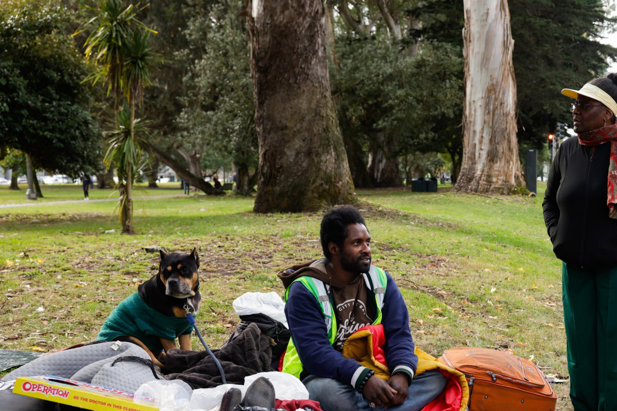 A man sits on the grass with belongings, including a skateboard and a suitcase. A dog in a sweater sits nearby, and a woman stands, facing him in conversation.