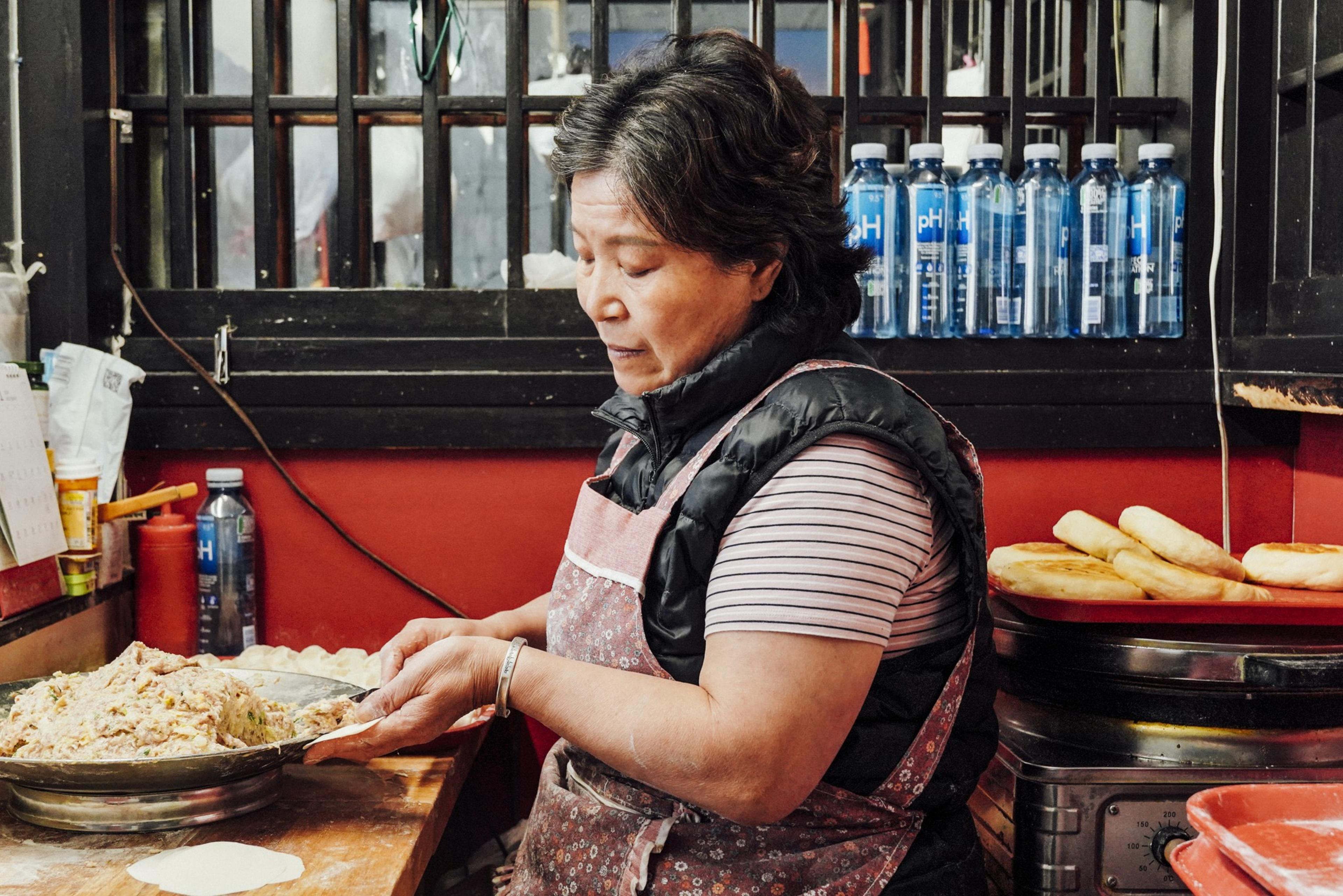 A woman stands in a kitchen preparing dough. She's wearing a striped shirt and apron, surrounded by a window, water bottles, and bread on a red tray.