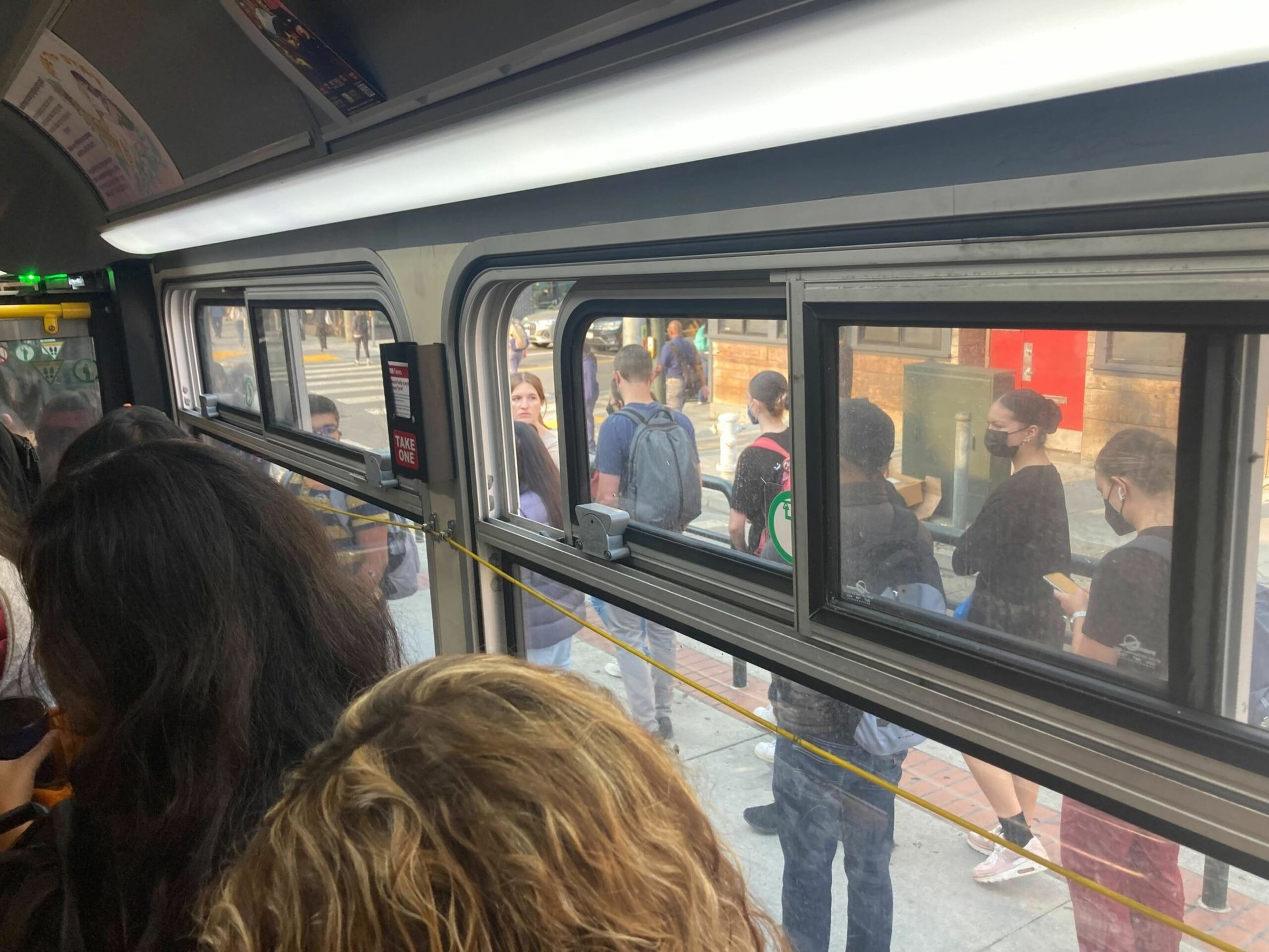 People stand inside a crowded bus while others wait outside at a bus stop, some wearing face masks and one person looking at their phone.